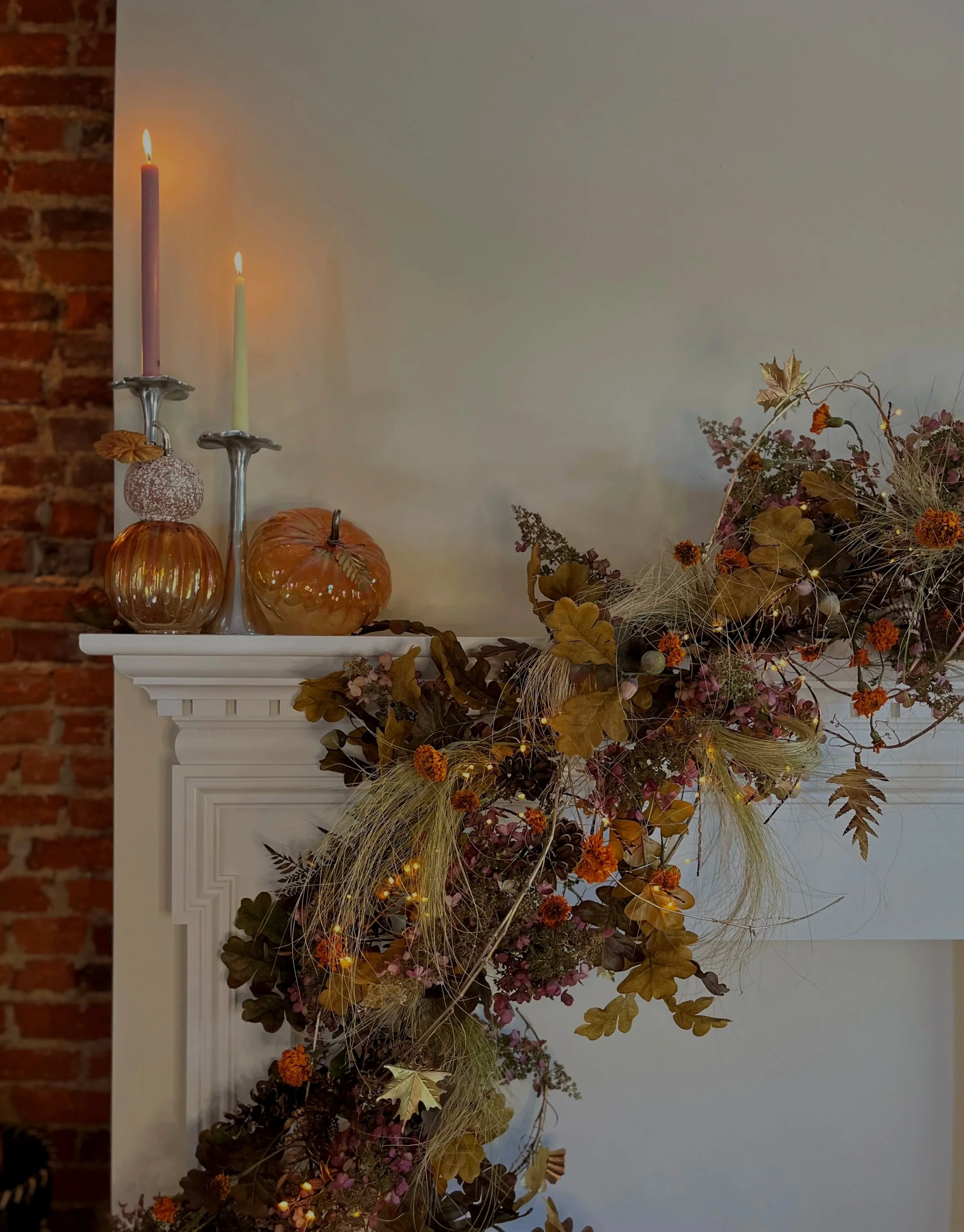 Autumn-themed decor including glass pumpkins, tall candlesticks with lit candles, and a floral arrangement with dried leaves, flowers, and grasses on a white mantelpiece against a white wall and brick wall background.