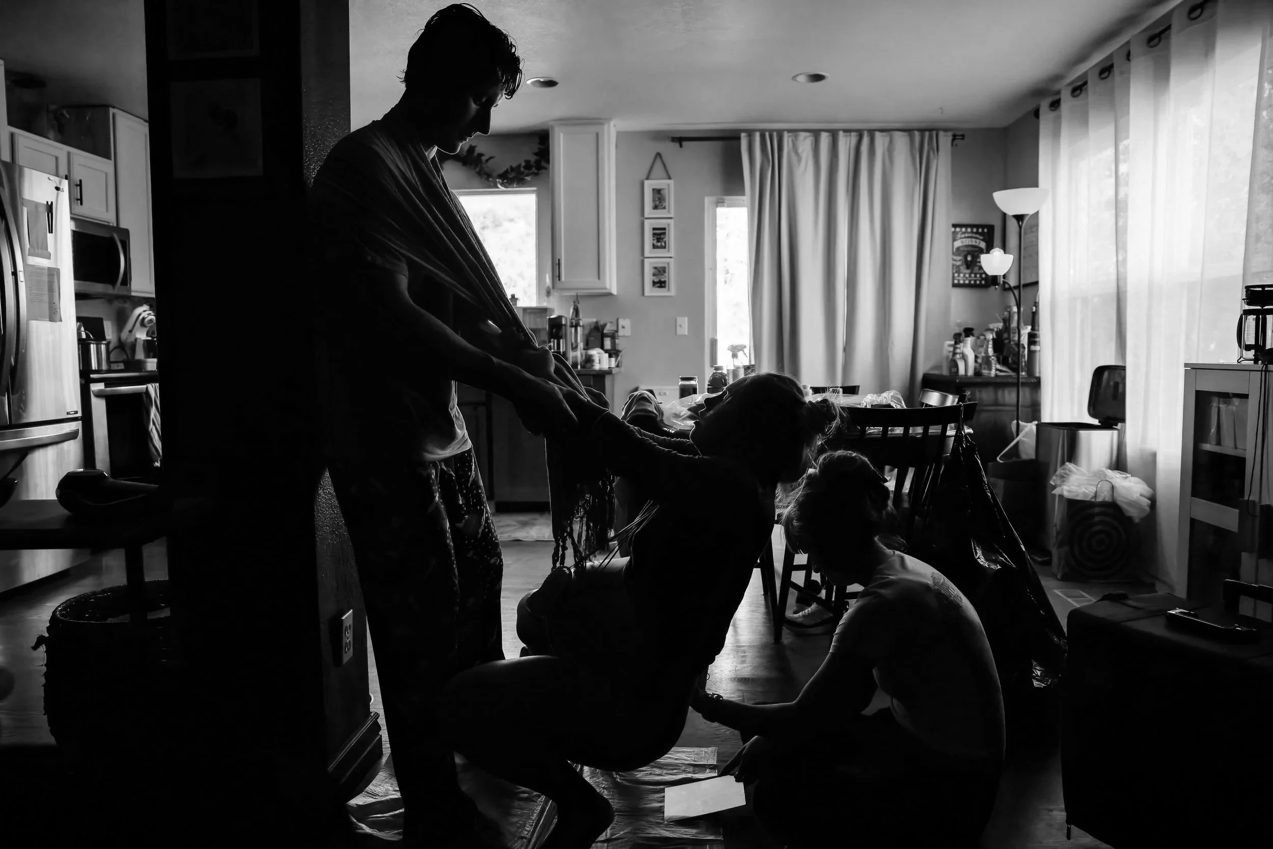 A person is standing and holding hands with a child sitting in a high chair in a kitchen filled with natural light, with curtains, framed pictures, and various household items visible.