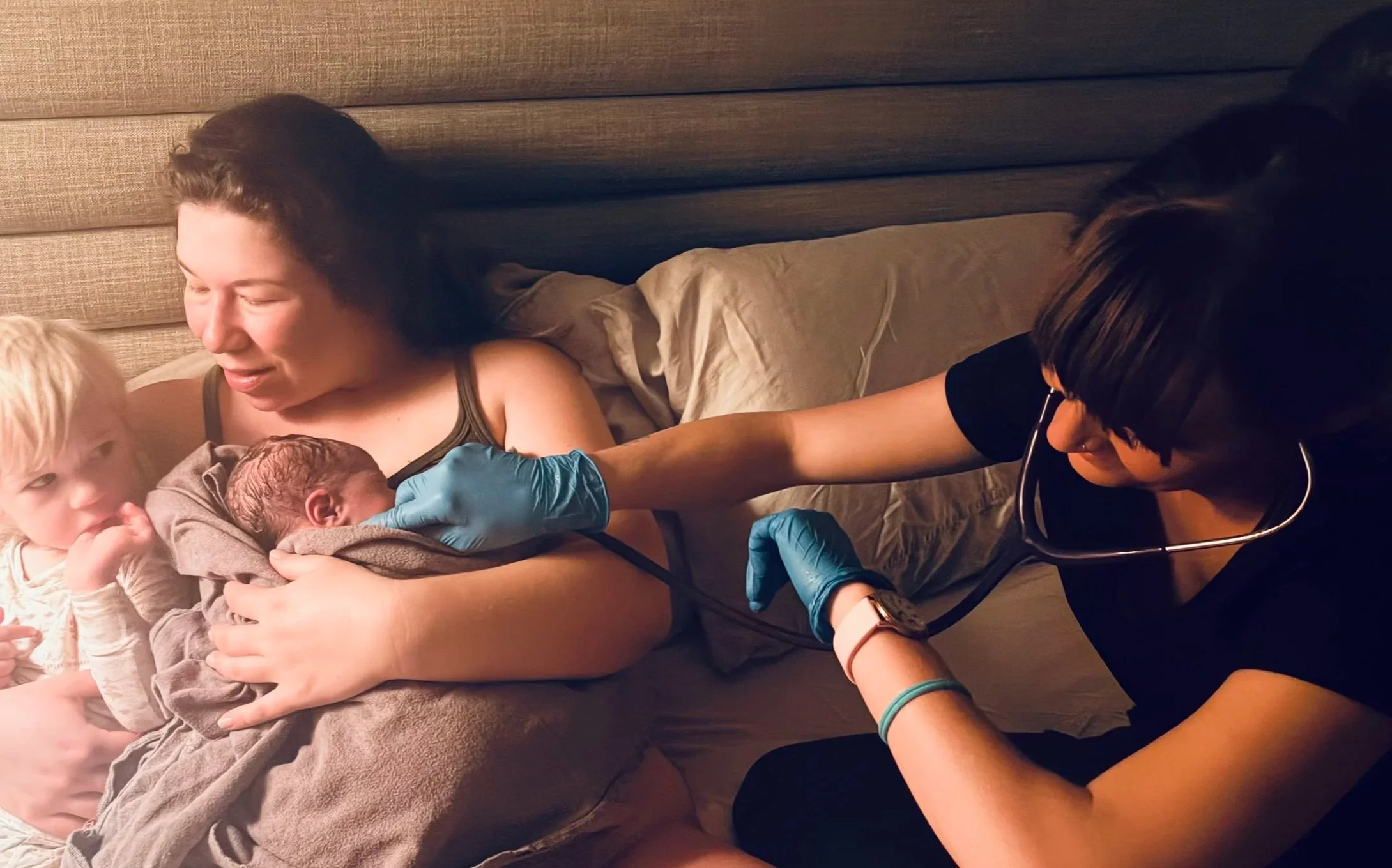 A woman holding a newborn baby with a toddler next to her, while a healthcare professional in scrubs and gloves checks the baby’s heartbeat with a stethoscope.