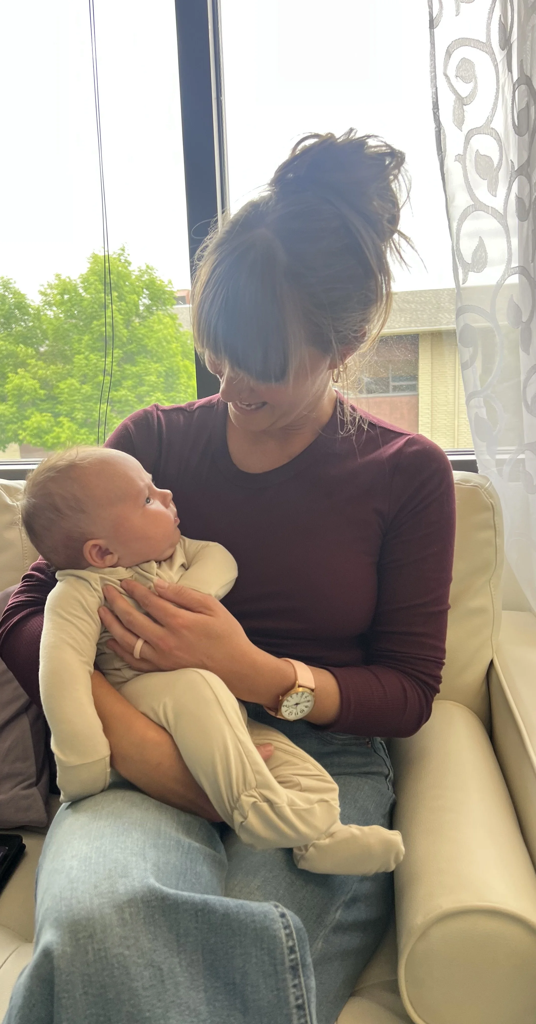 A woman with a bun hairstyle, wearing a maroon long sleeve shirt and a watch, is sitting on a beige chair holding a baby. The baby is looking up at the woman and is wearing a cream-colored outfit. They are sitting near a window with white curtains, and outside there are green trees and part of a building.
