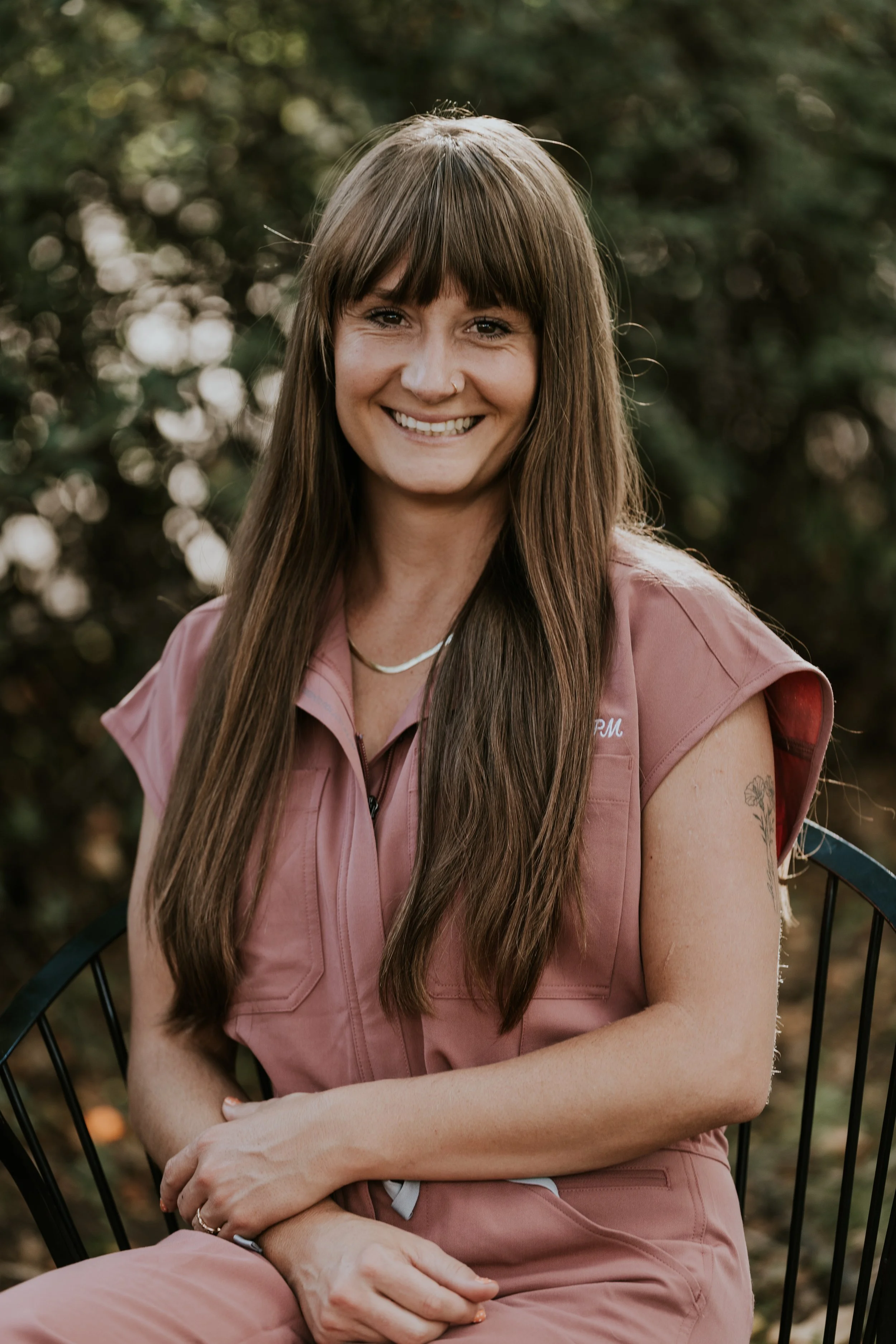 A woman with long brown hair and a nose piercing, smiling, sitting outdoors on a black wire chair, wearing a pink sleeveless vest and pants, with trees in the background.