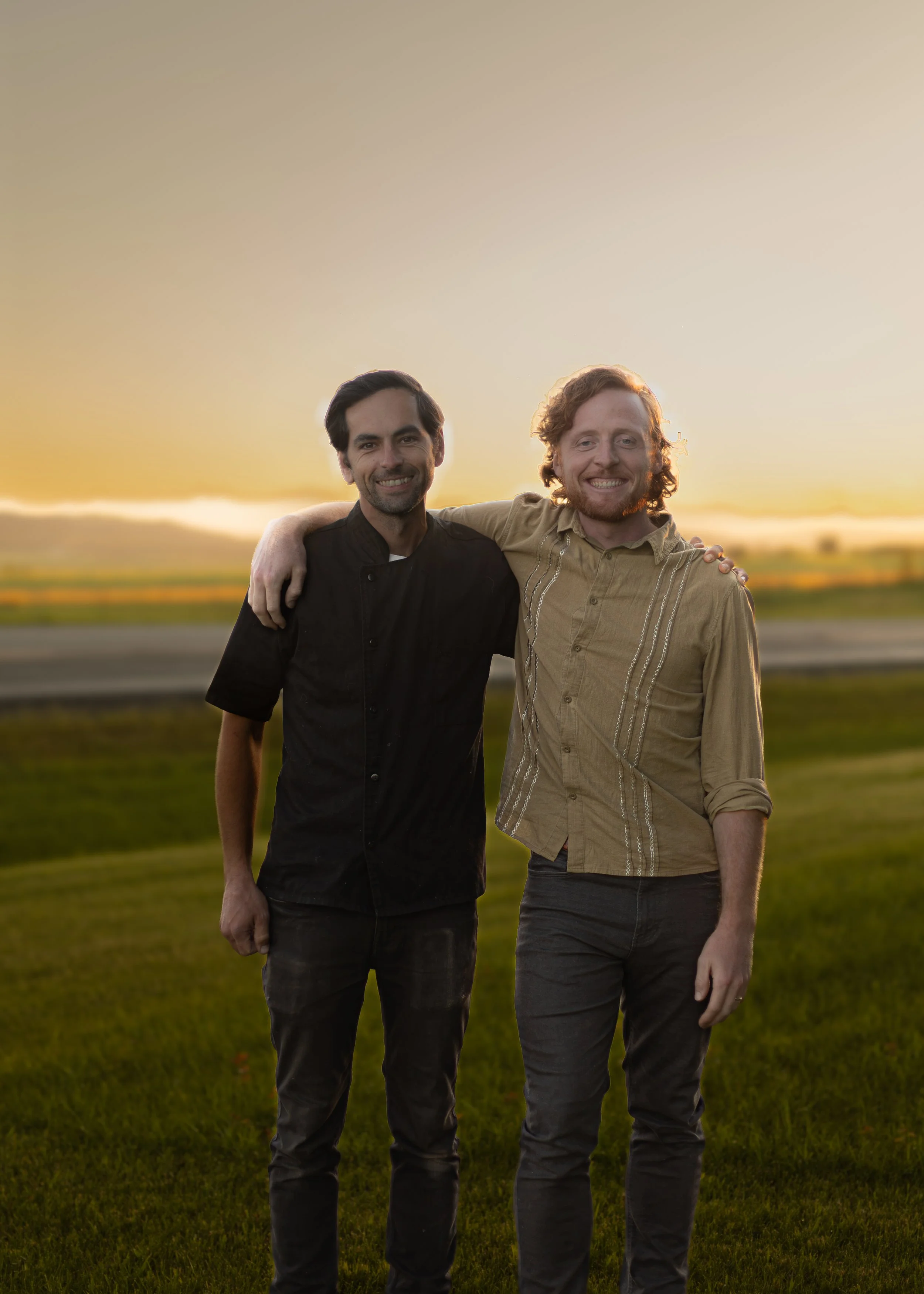 Two men standing outdoors on grass at sunset, smiling with arms around each other.
