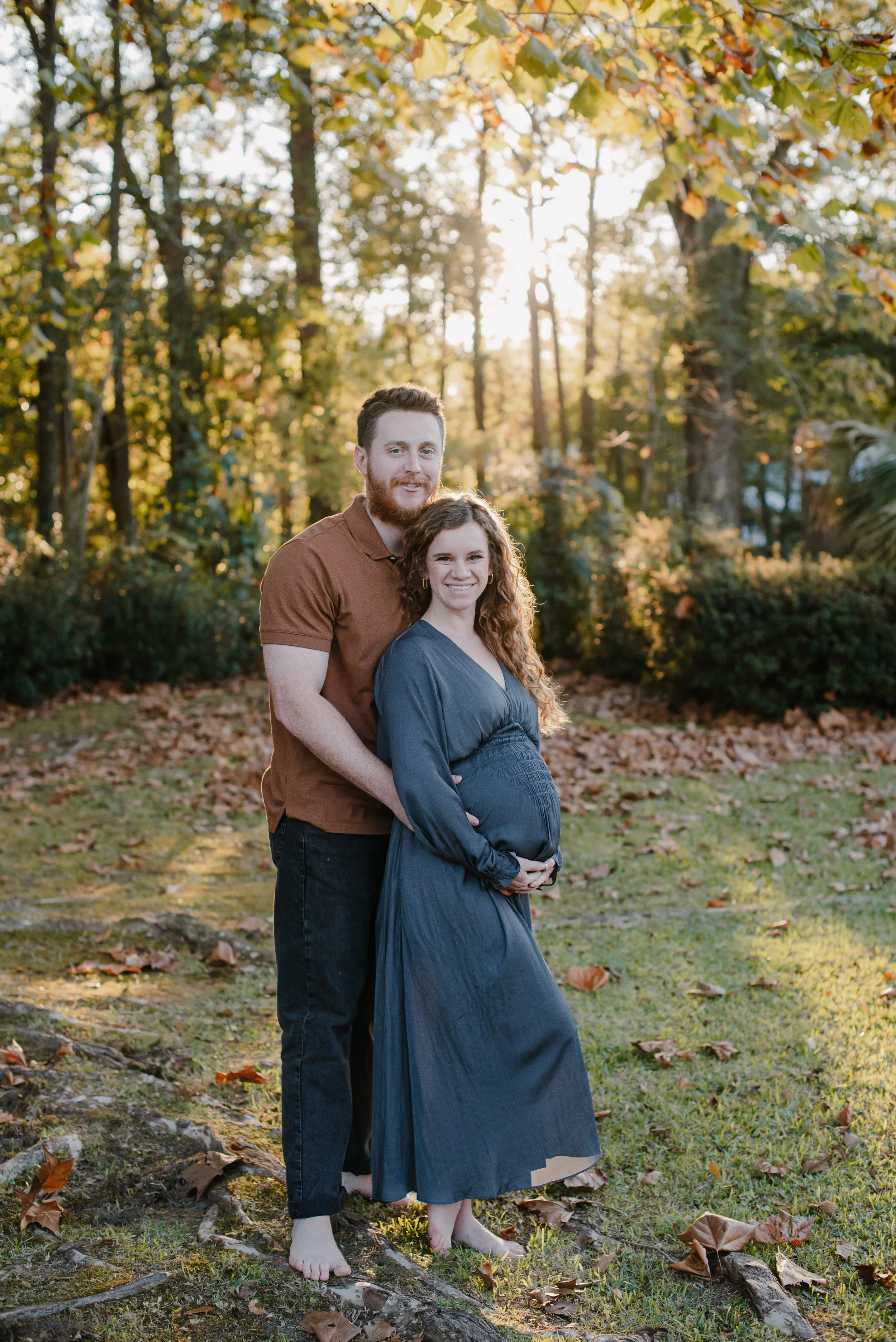 A pregnant woman in a long dress and barefoot stands in front of a man in an orange shirt and black jeans in a wooded area during autumn. They are smiling and standing close together, with some fallen leaves on the ground and sunlight filtering through the trees.