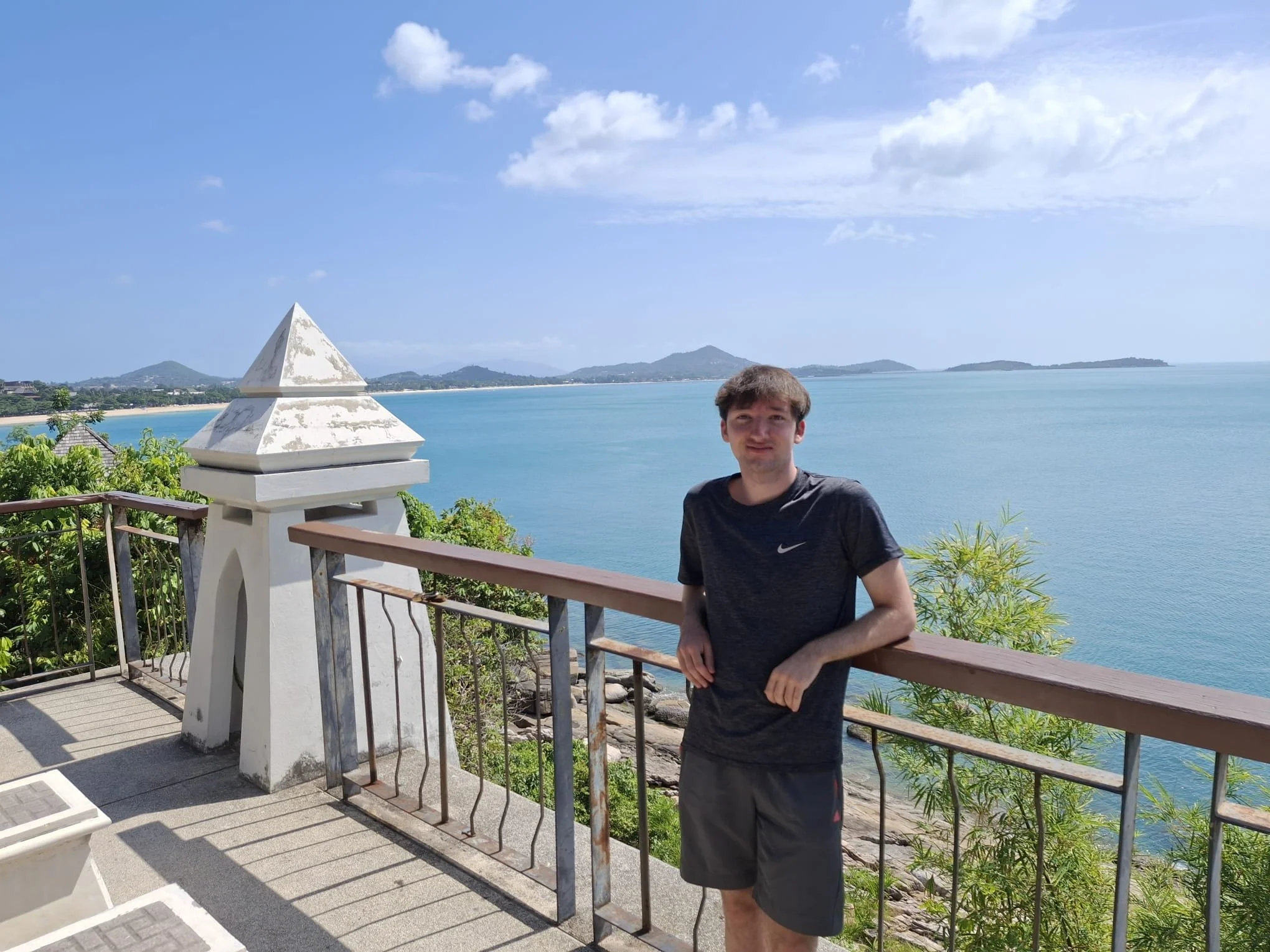 A young man standing on a balcony with a view of a blue ocean, distant mountains, and a partly cloudy sky. He is wearing a black Nike t-shirt and black shorts.