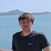 A young man standing outdoors near water with mountains in the background, wearing a dark Nike t-shirt.