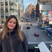 Young woman with long hair smiling, standing outdoors in an urban city with tall buildings and busy street in the background.