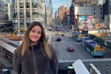 A young woman with long brown hair smiling in an urban cityscape with tall buildings, yellow taxis, and buses on the street.