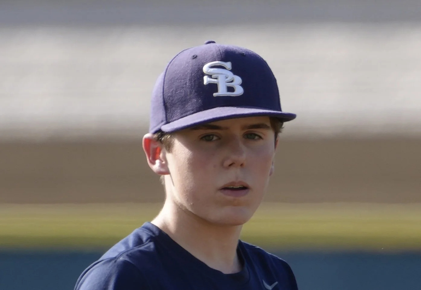 Close-up of a young male baseball player with a serious expression, wearing a navy blue cap with 'SB' logo, and a navy shirt, outdoors