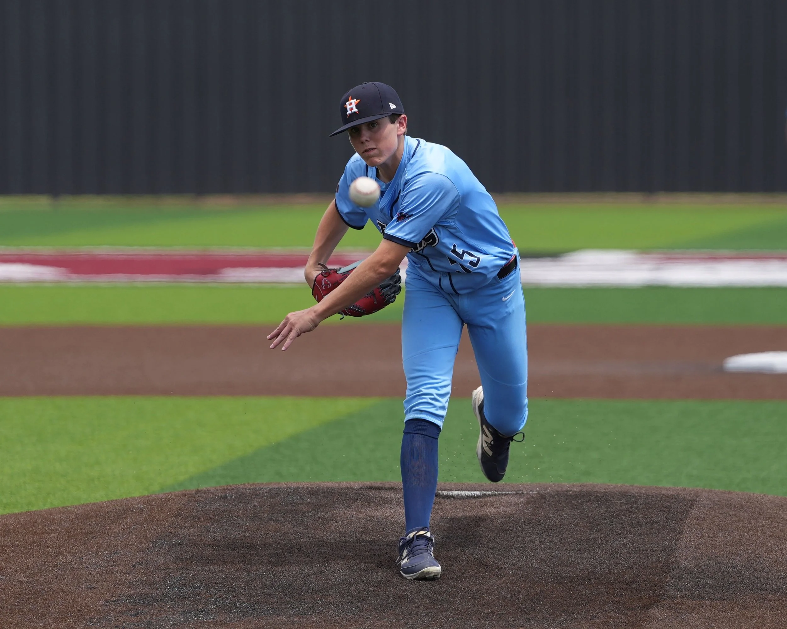 A young baseball player in a light blue uniform and black cap on a baseball field, about to pitch a ball.