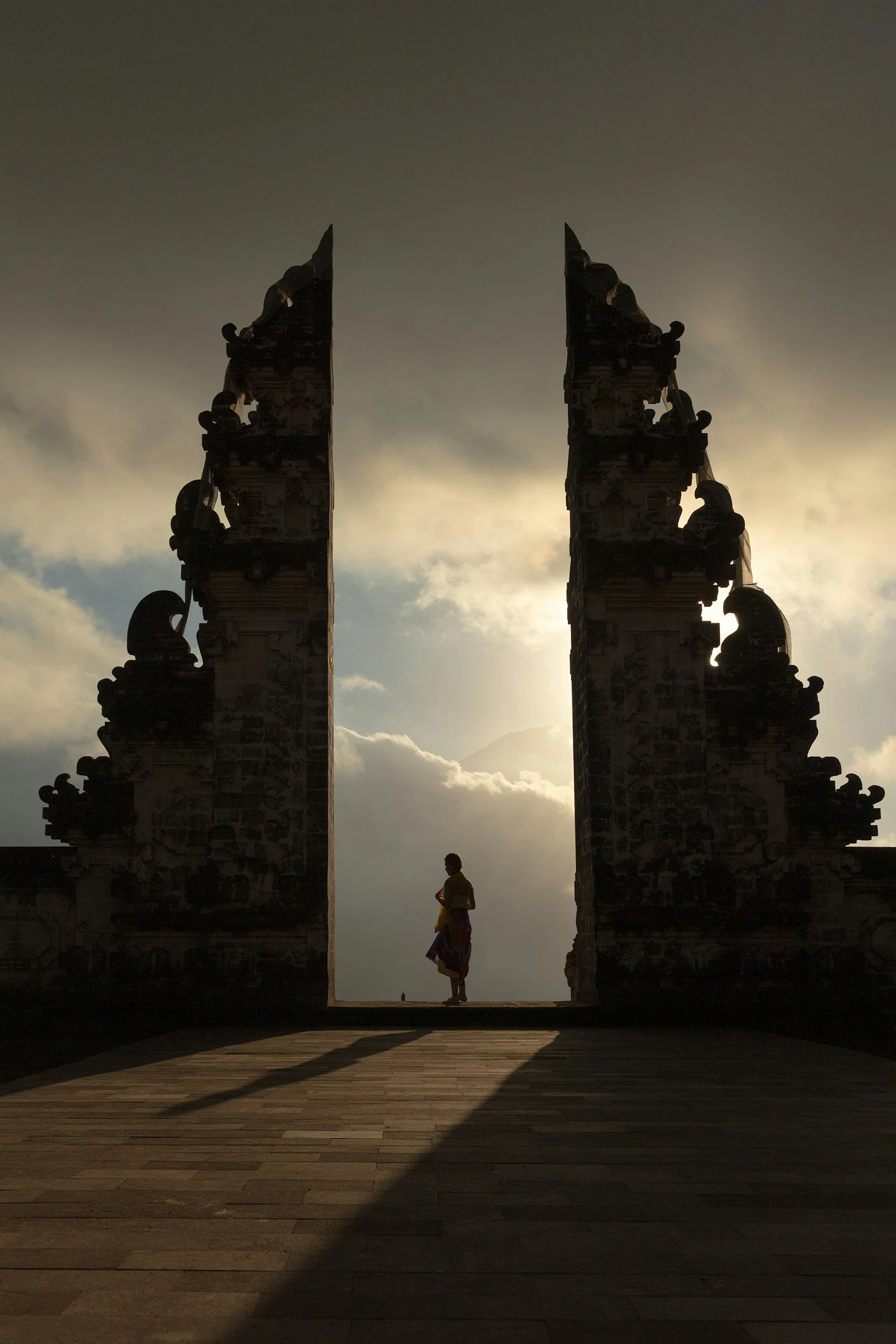 Silhouette of a person standing in front of a large, ornate gateway with intricate carvings, set against a cloudy sky with the sun near the horizon.