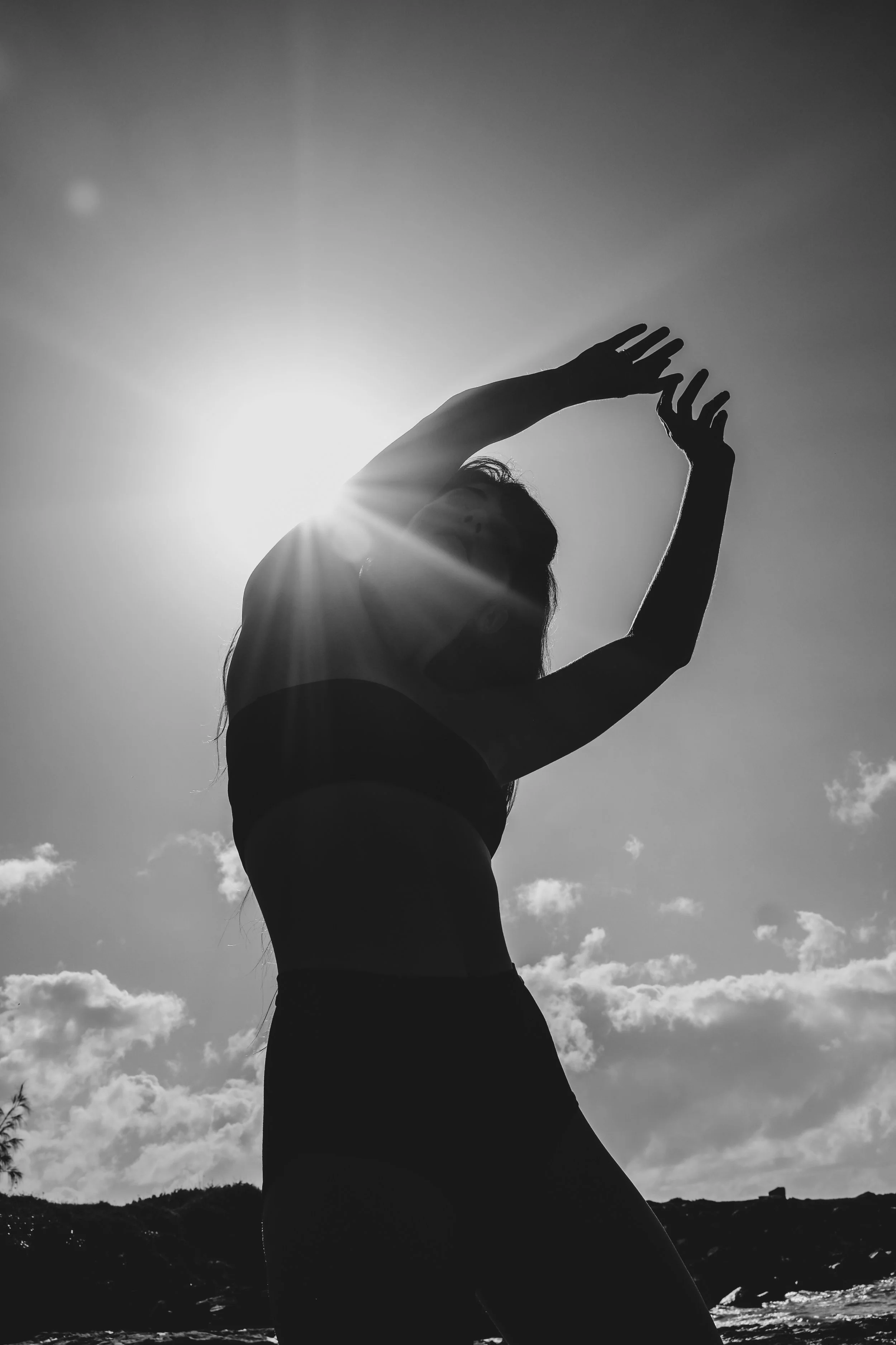 A silhouette of a woman standing outdoors against a bright sun and cloudy sky, with distant hills in the background.