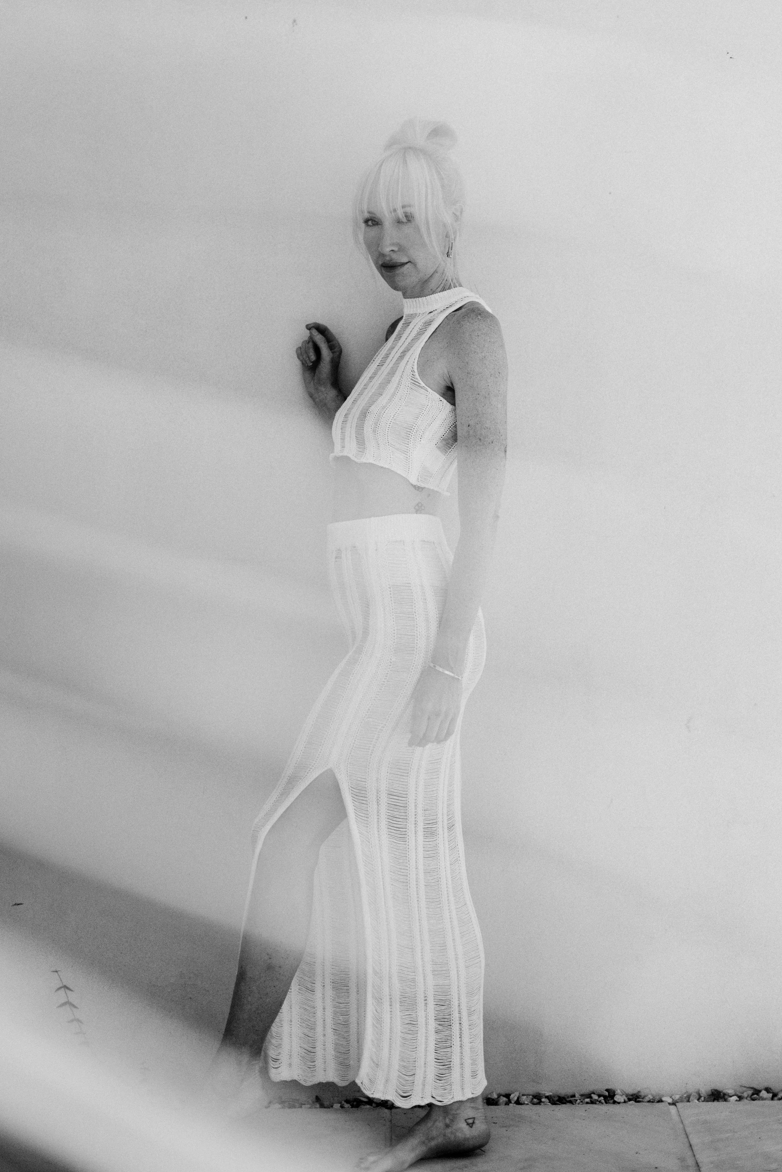 A woman with blond hair tied in a bun, wearing a sleeveless knitted top and striped pants, standing against a plain wall, looking at the camera, with a tattoo on her foot.