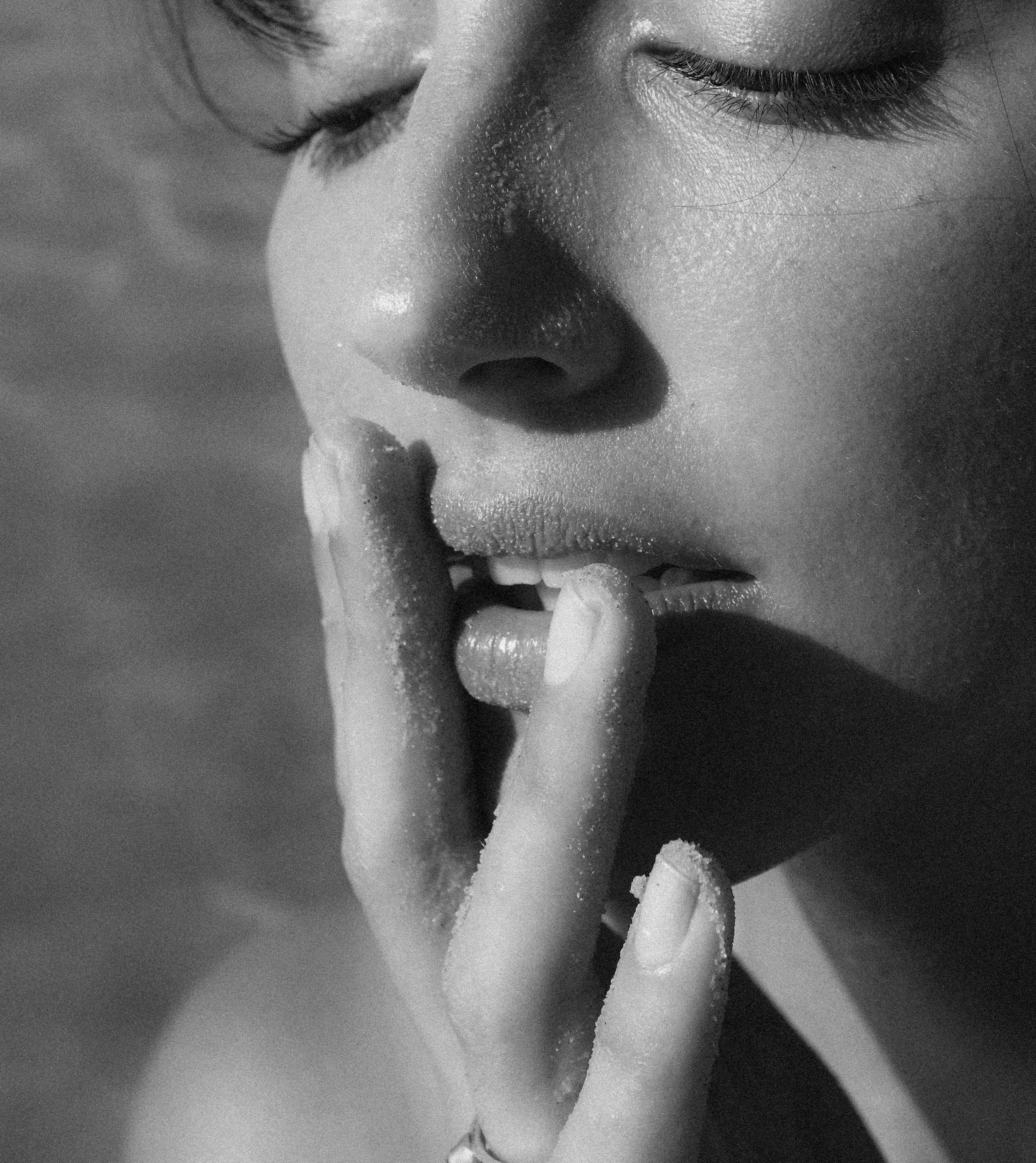Close-up black and white photograph of a woman with her eyes closed and finger touching her lips, emphasizing her beautiful skin and features.