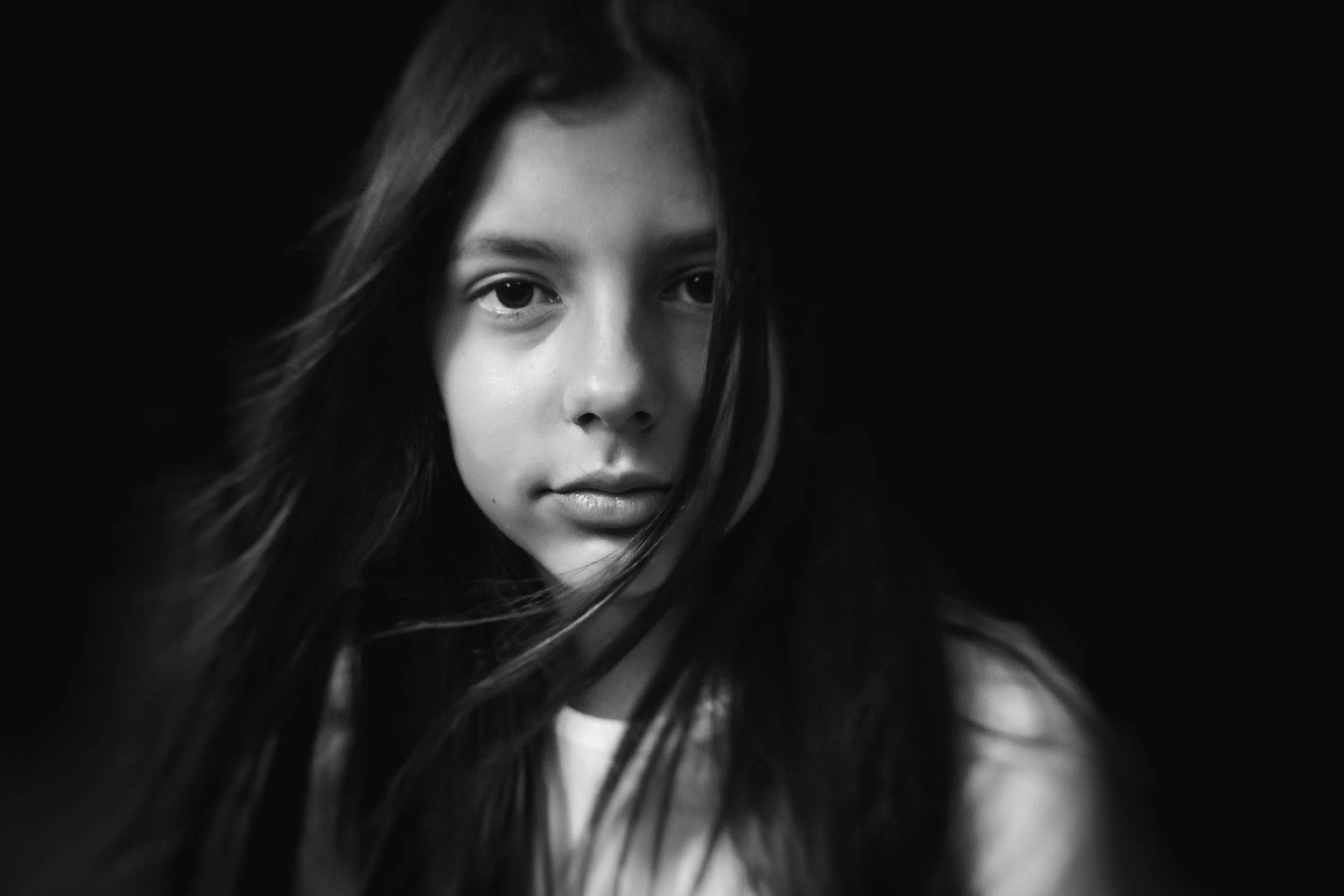 A black-and-white close-up portrait of a woman with long hair, looking directly at the camera with a serious expression, partially obscured by dark surroundings.