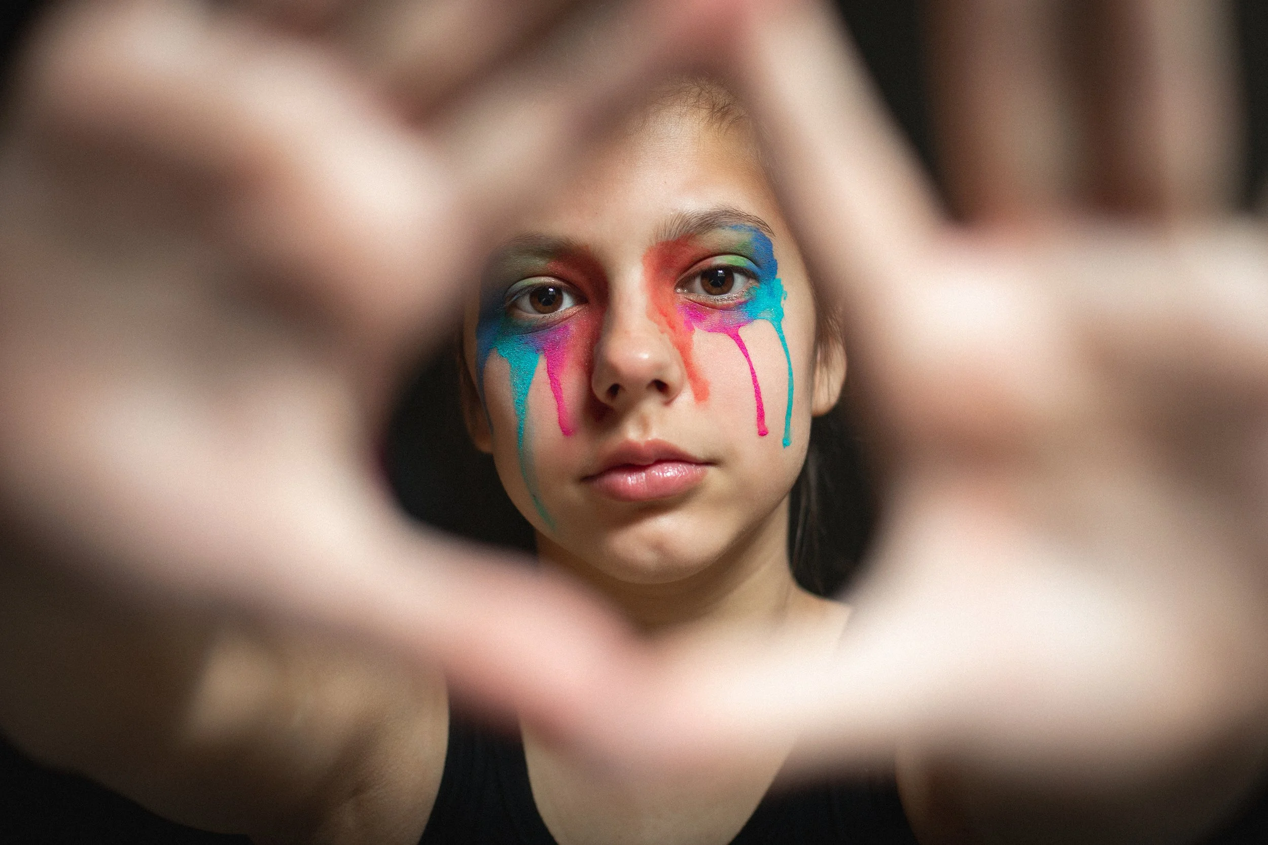 A young person with rainbow-colored paint dripping down their face, framed by blurred fingers.