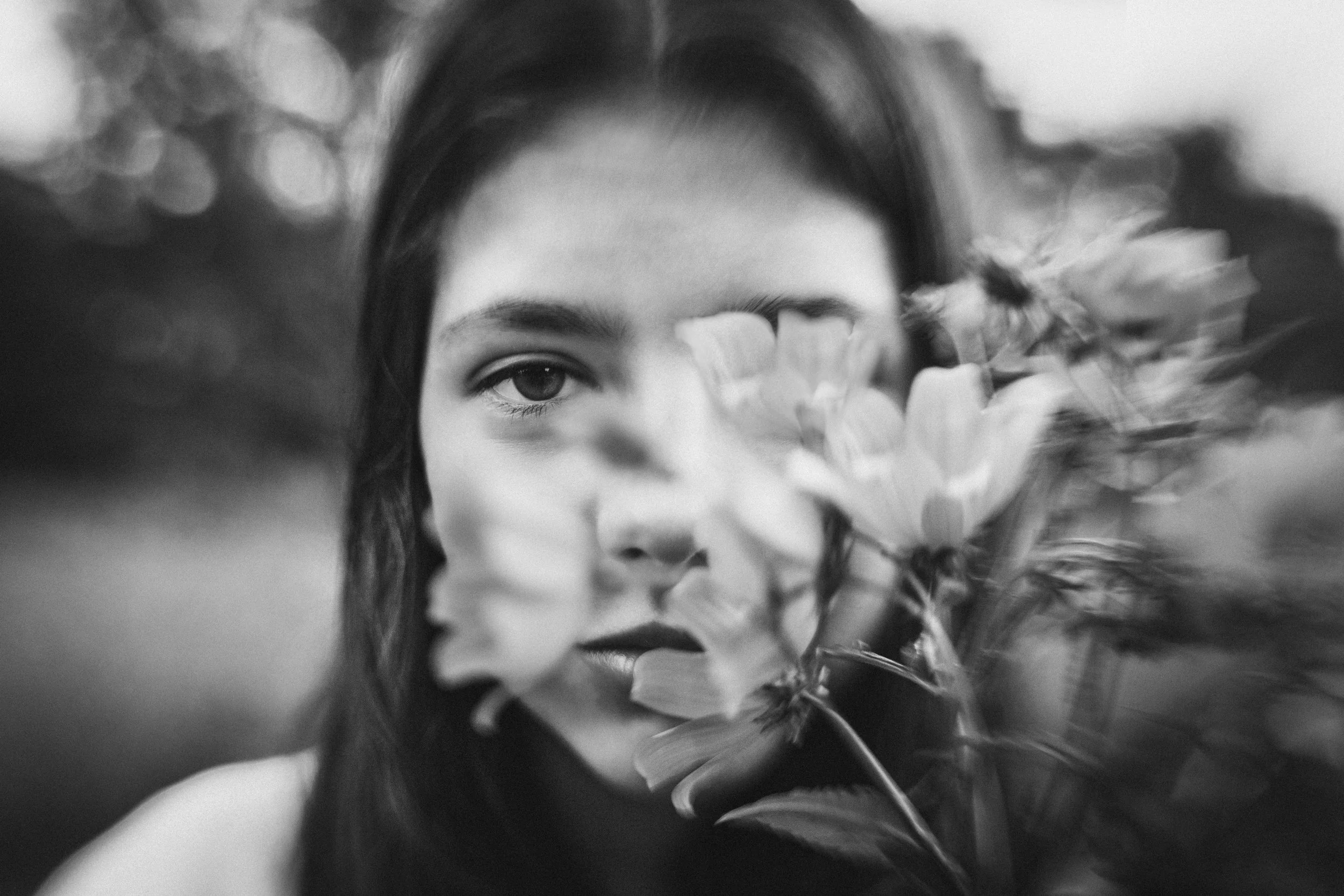 Black and white photograph of a woman with dark hair, partially obscured by large flowers in front of her face, one eye and part of her lips visible.