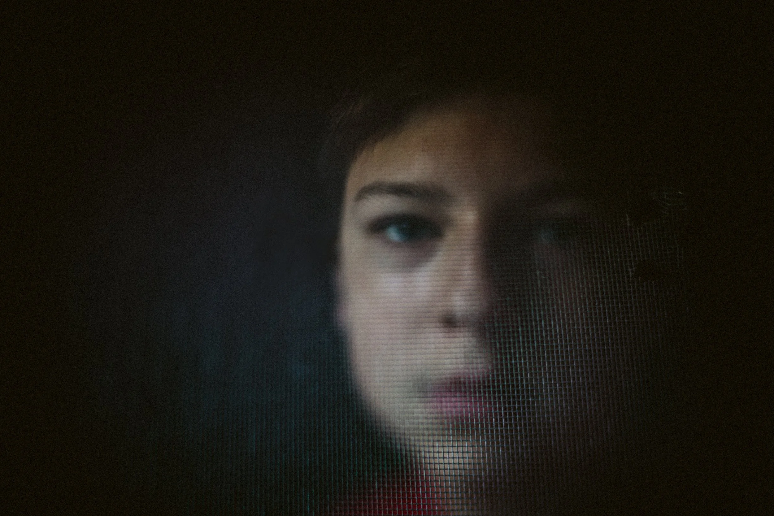 Close-up of a person's face behind a mesh or screen, with one eye and part of the face visible.