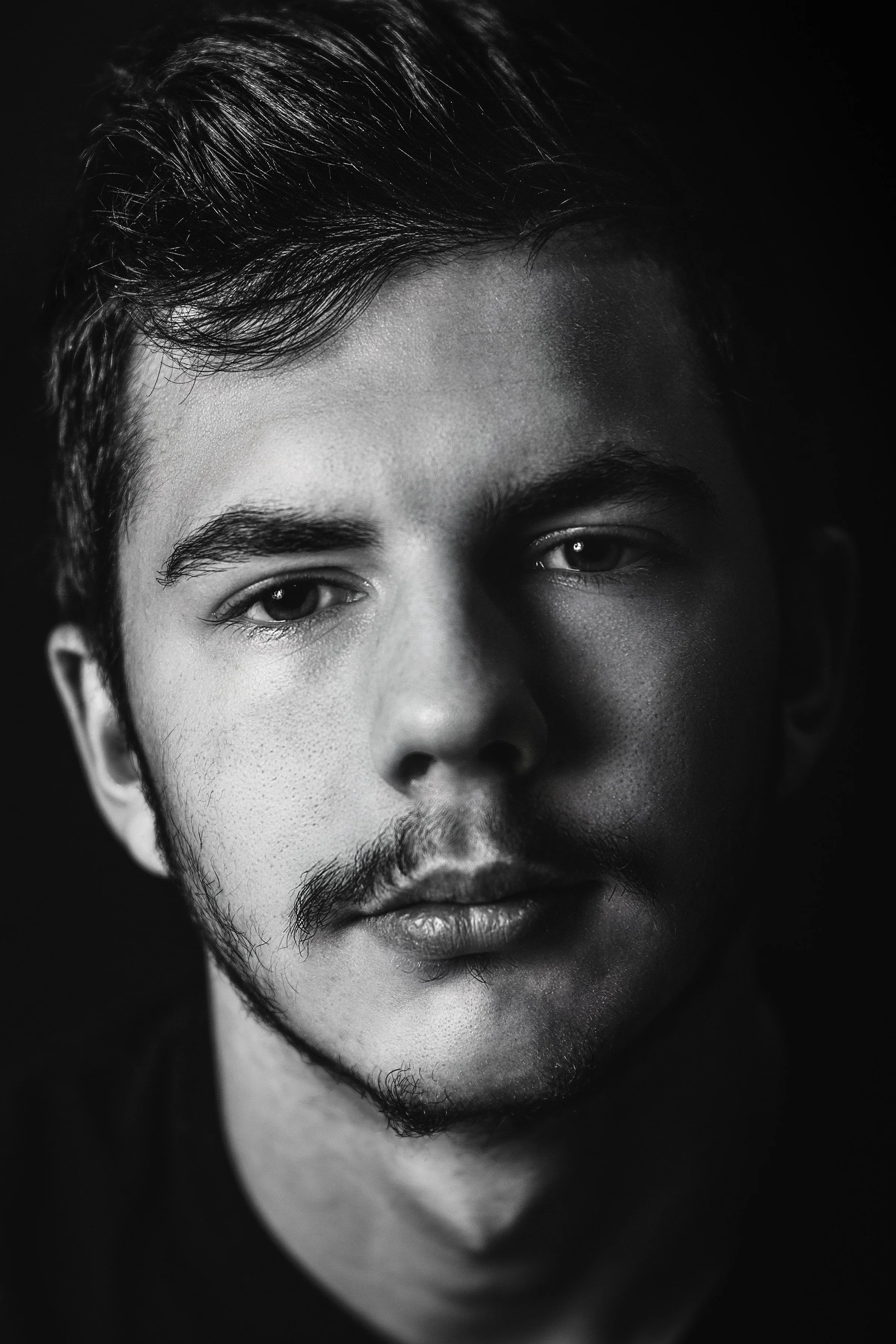 Close-up black and white portrait of a young man's face showing his eyes, forehead, nose, lips, and beard, with a dark background.