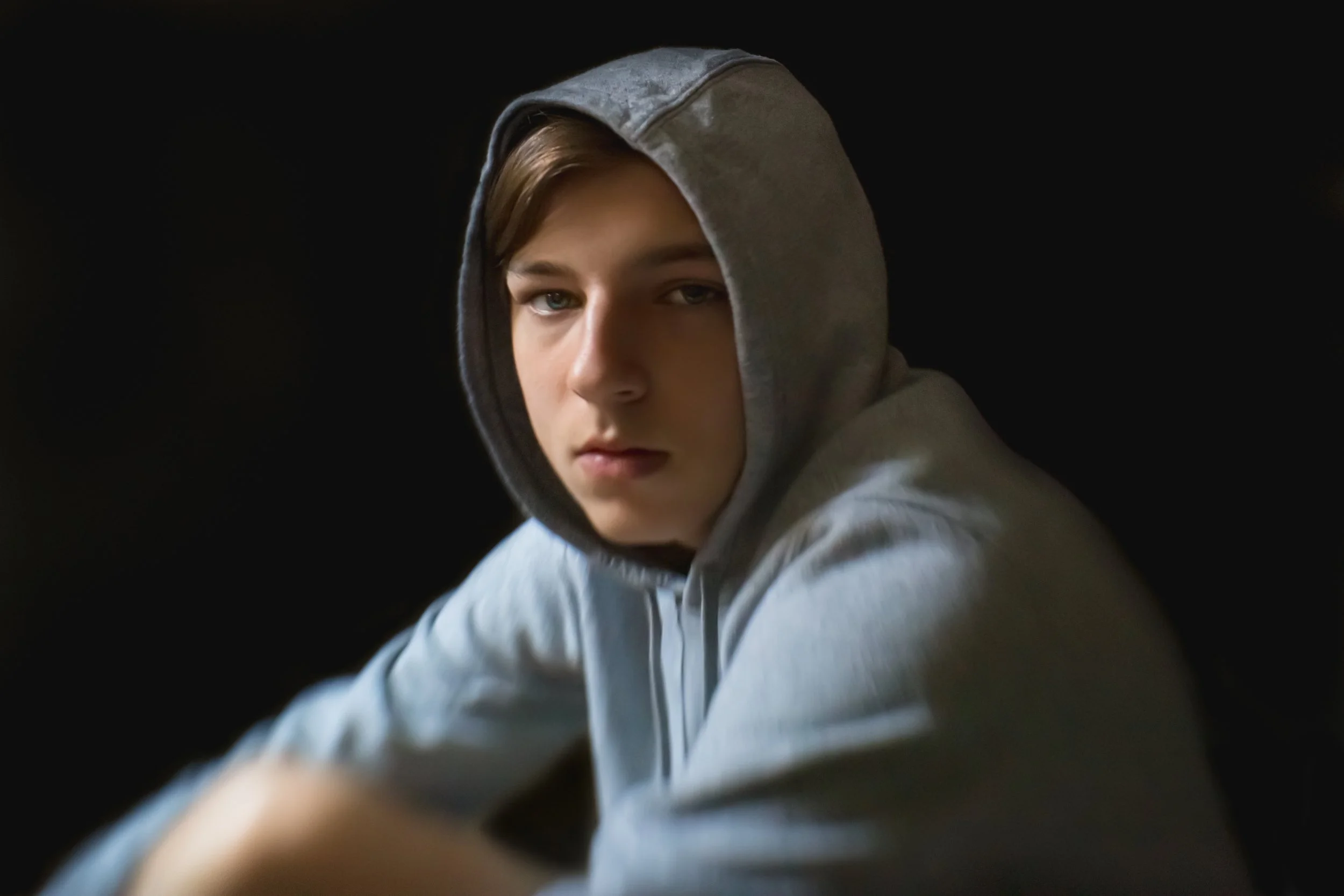 A young person with light skin and blue eyes wearing a gray hoodie, looking slightly to the side against a black background.