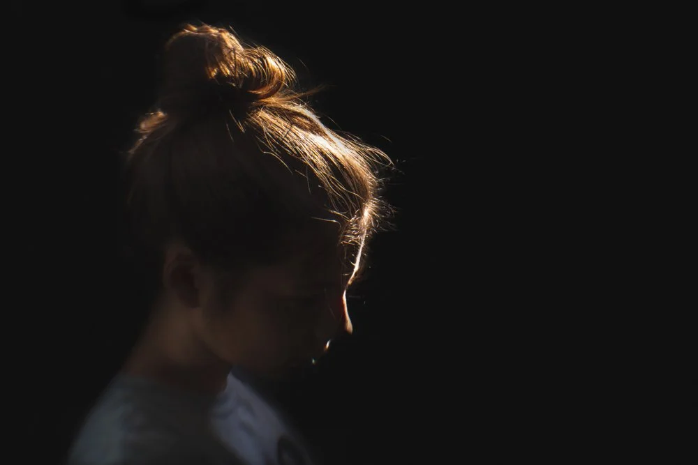 Side profile of a woman with her hair in a bun, illuminated by a rim light  in a dark background.