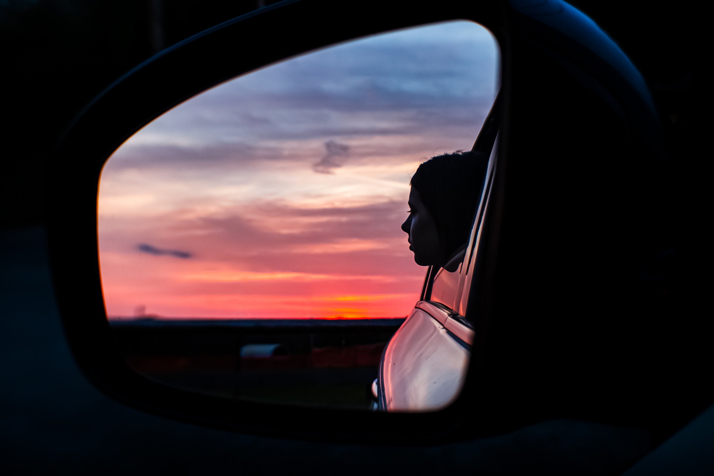 A woman with dark hair looking out of a car window at a colorful sunset sky.