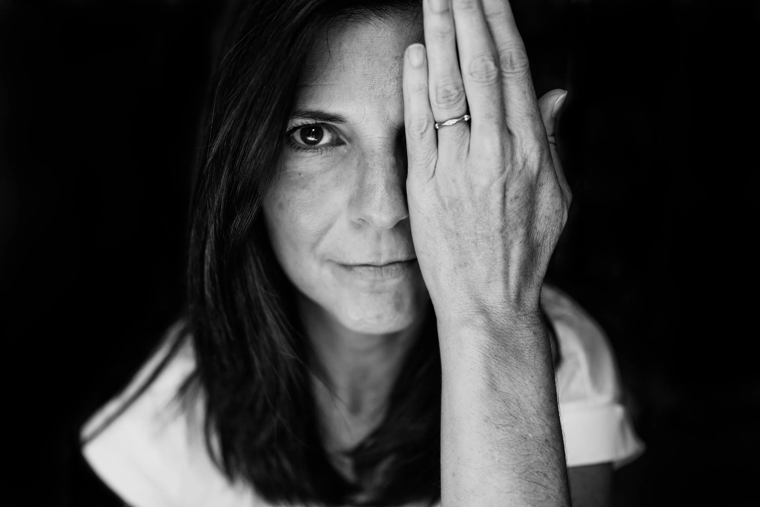 A black and white photo of a woman with dark hair, covering part of her face with her hand, showing only one eye and part of her face.