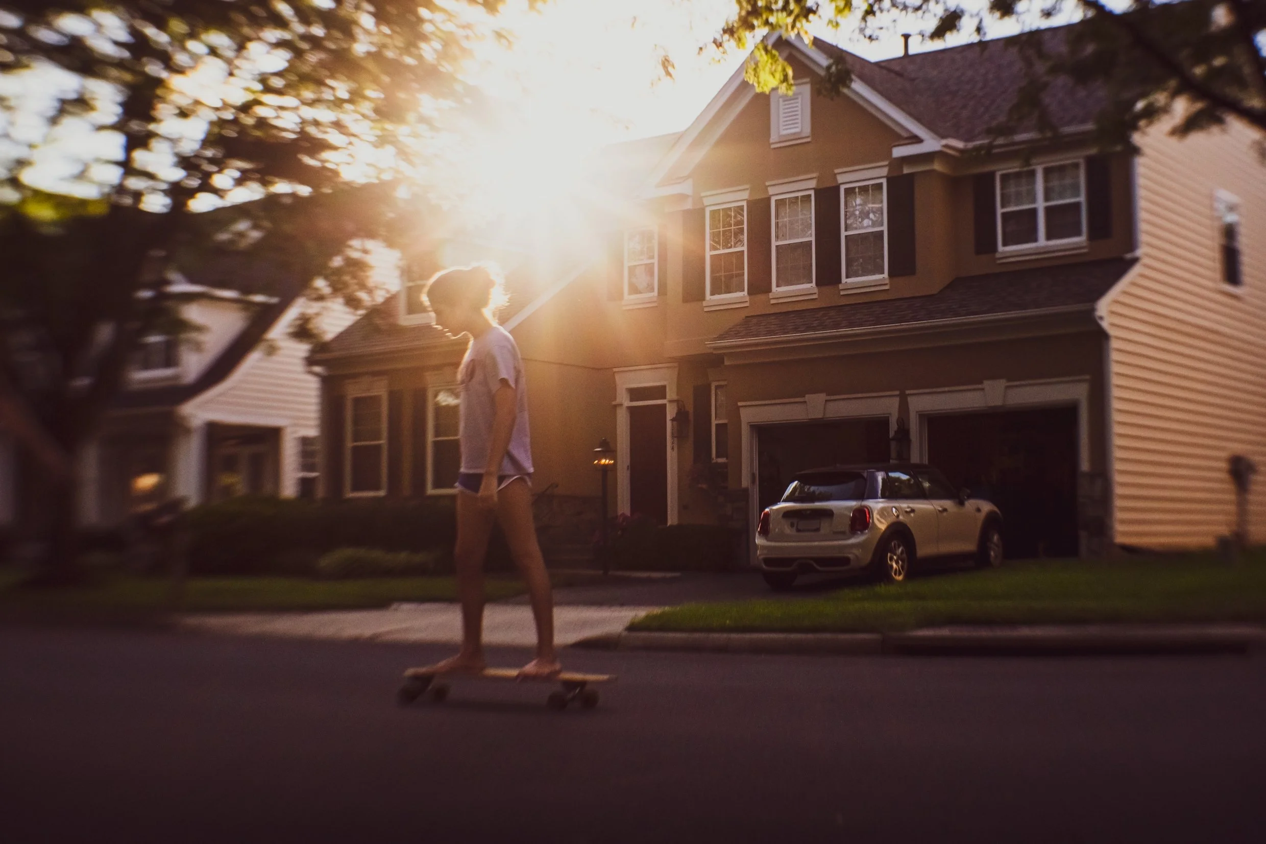 A person skateboarding on a suburban street at sunset, with houses, trees, and a parked car visible in the background.