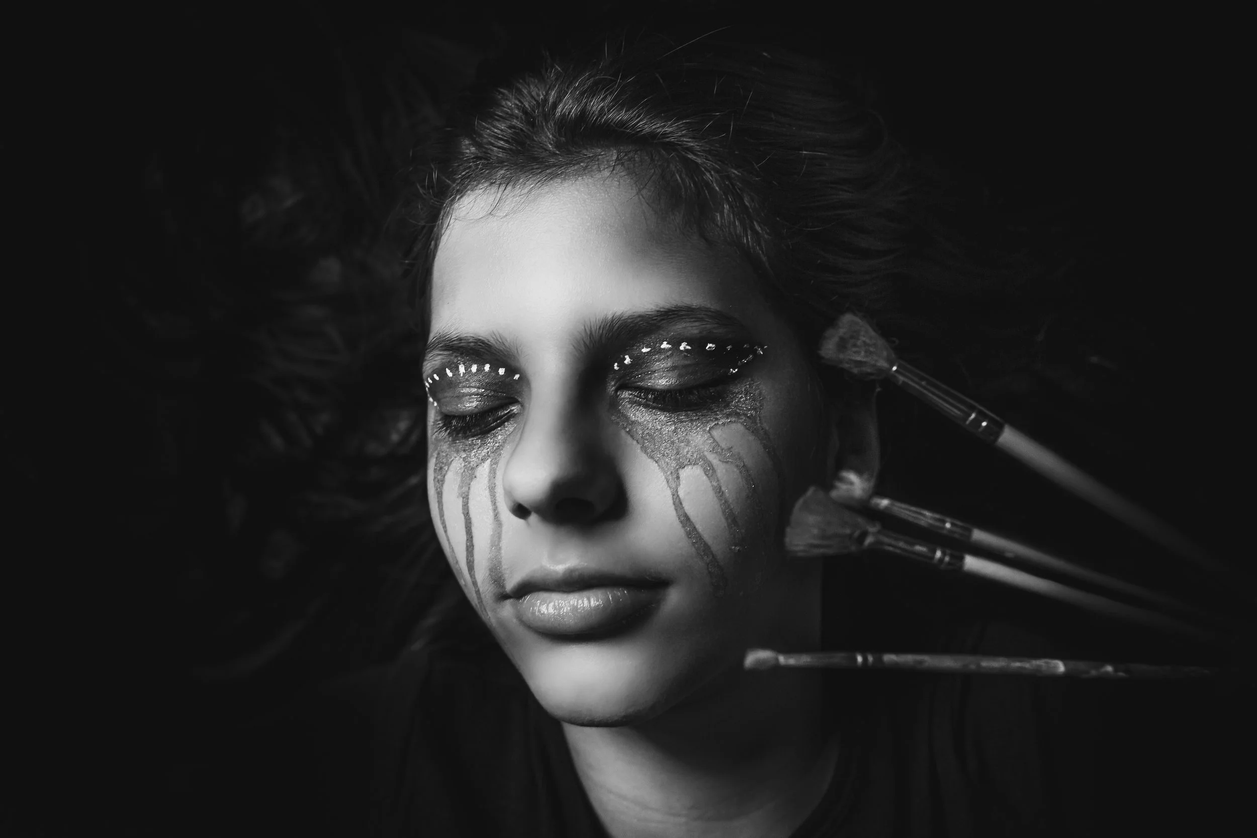 A woman with makeup, with brushes in her hand, her face partially painted or decorated with artistic designs, in black and white.