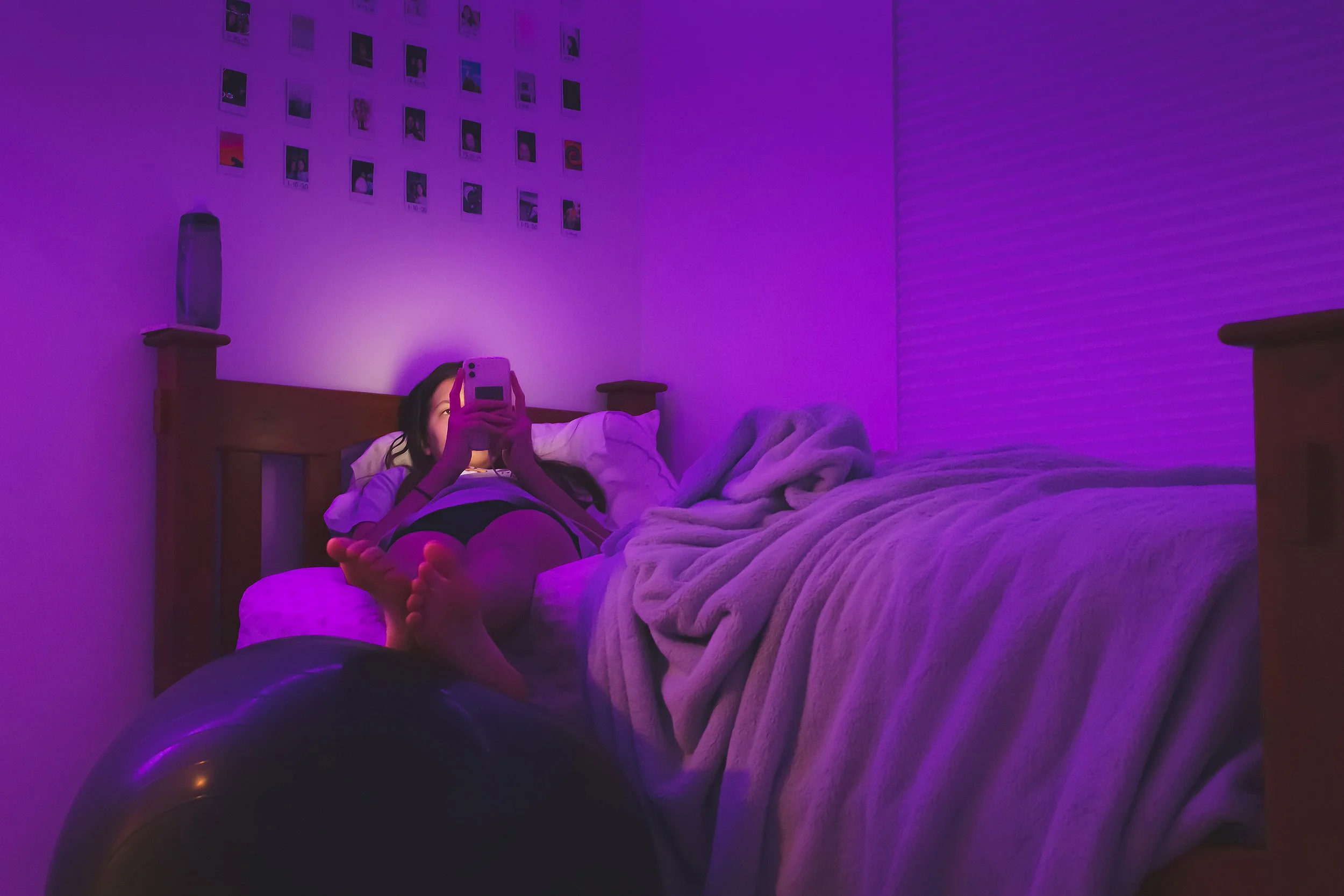 A person lying on a bed under purple/blue lighting, holding a smartphone in front of their face, with their feet visible. The bed has a blanket and pillows, and there are photo prints on the wall above the bed.