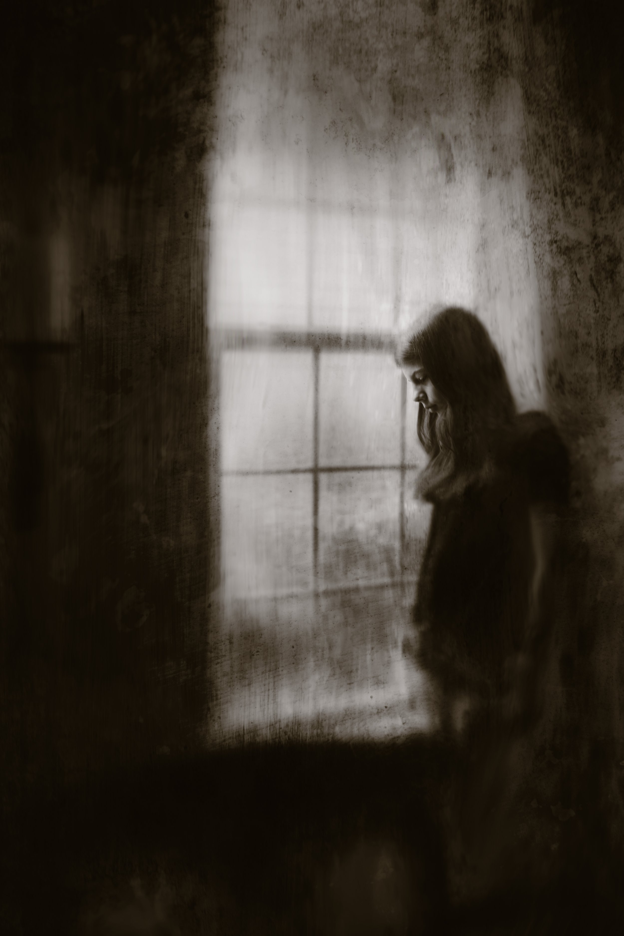 A person with long hair, appearing to be a young woman, is standing and looking downward in a dimly lit room with textured walls and a window in the background.