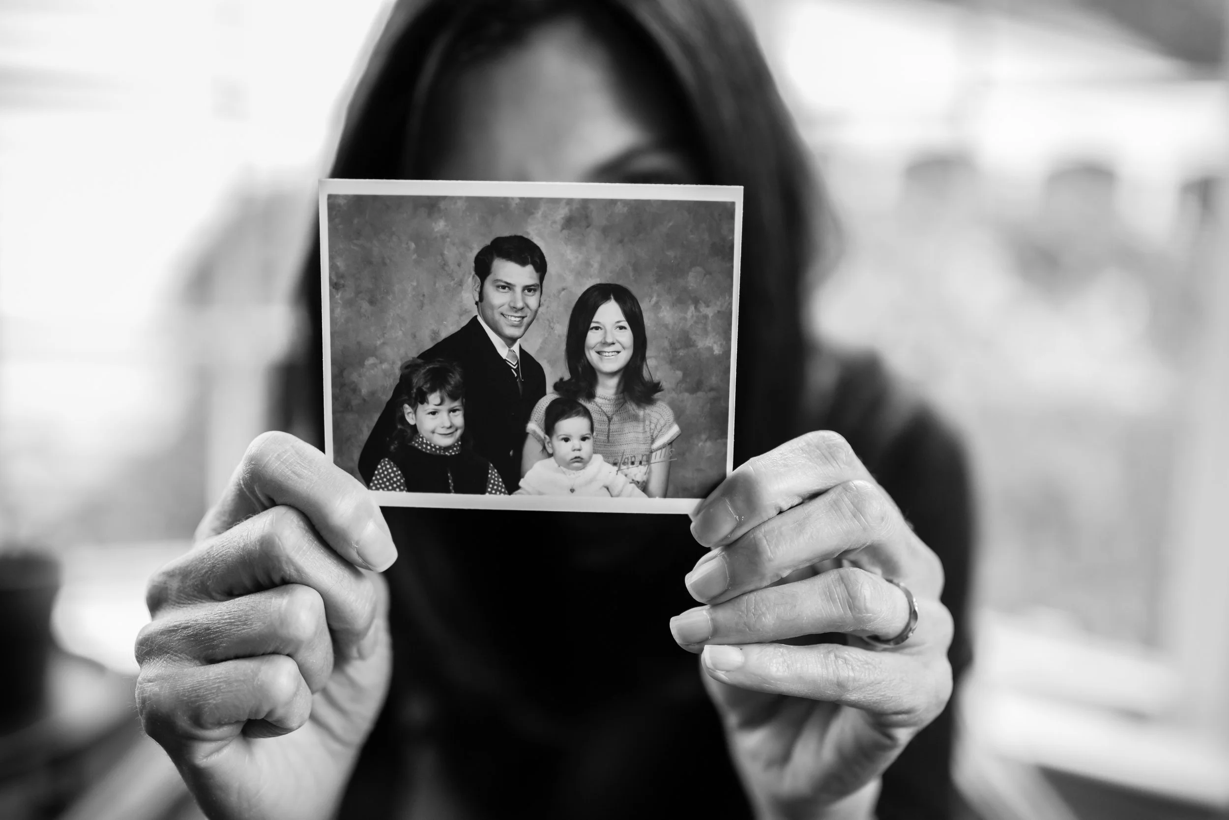A person holding a black and white photograph of a family portrait. The hands are visible, and the face of the person holding the photo is partially obscured, with only part of the forehead and hair showing.