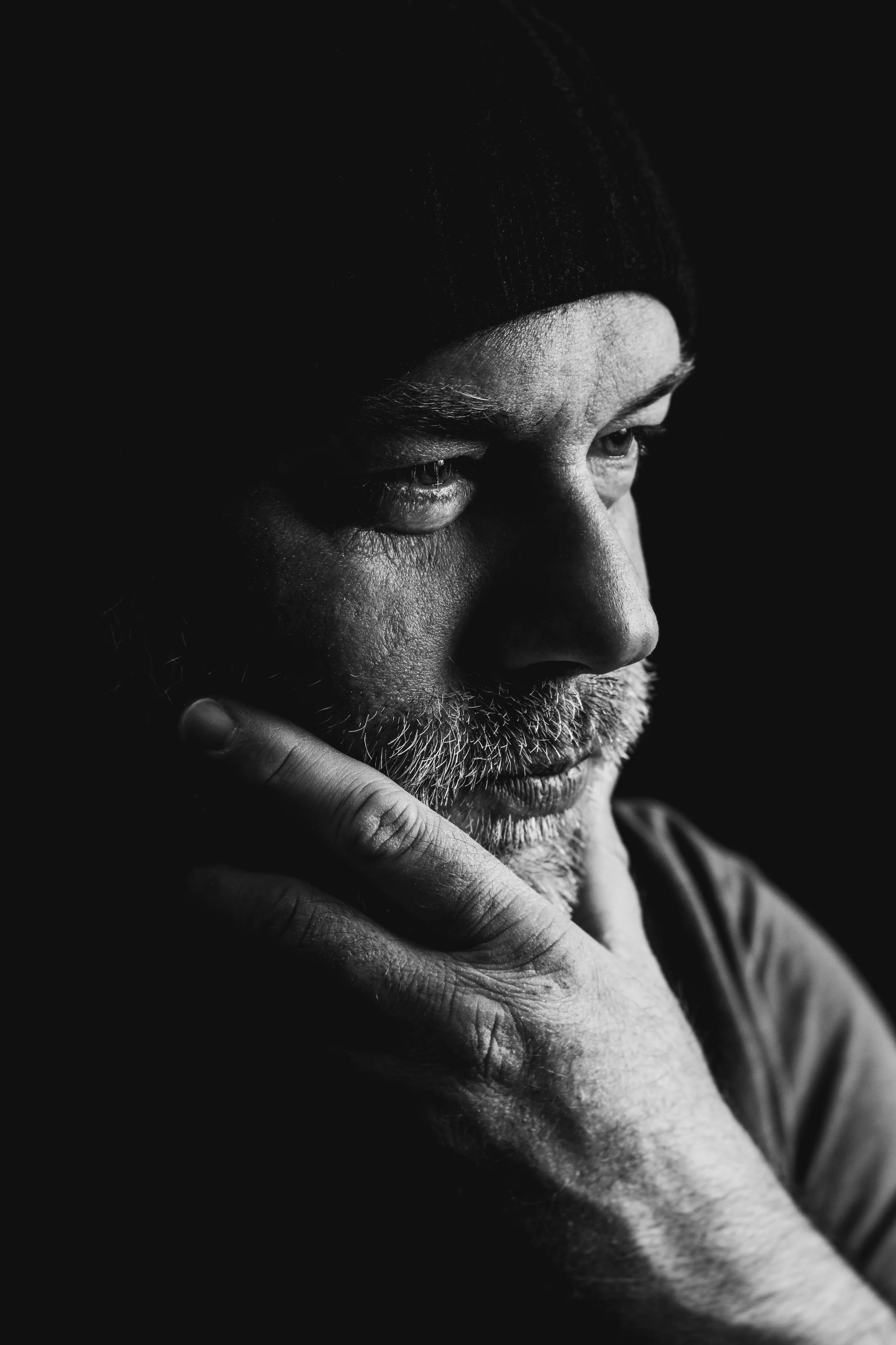 Close-up black and white photo of an elderly man with a beard, wearing a beanie, looking thoughtfully downward, with his hand resting on his chin.