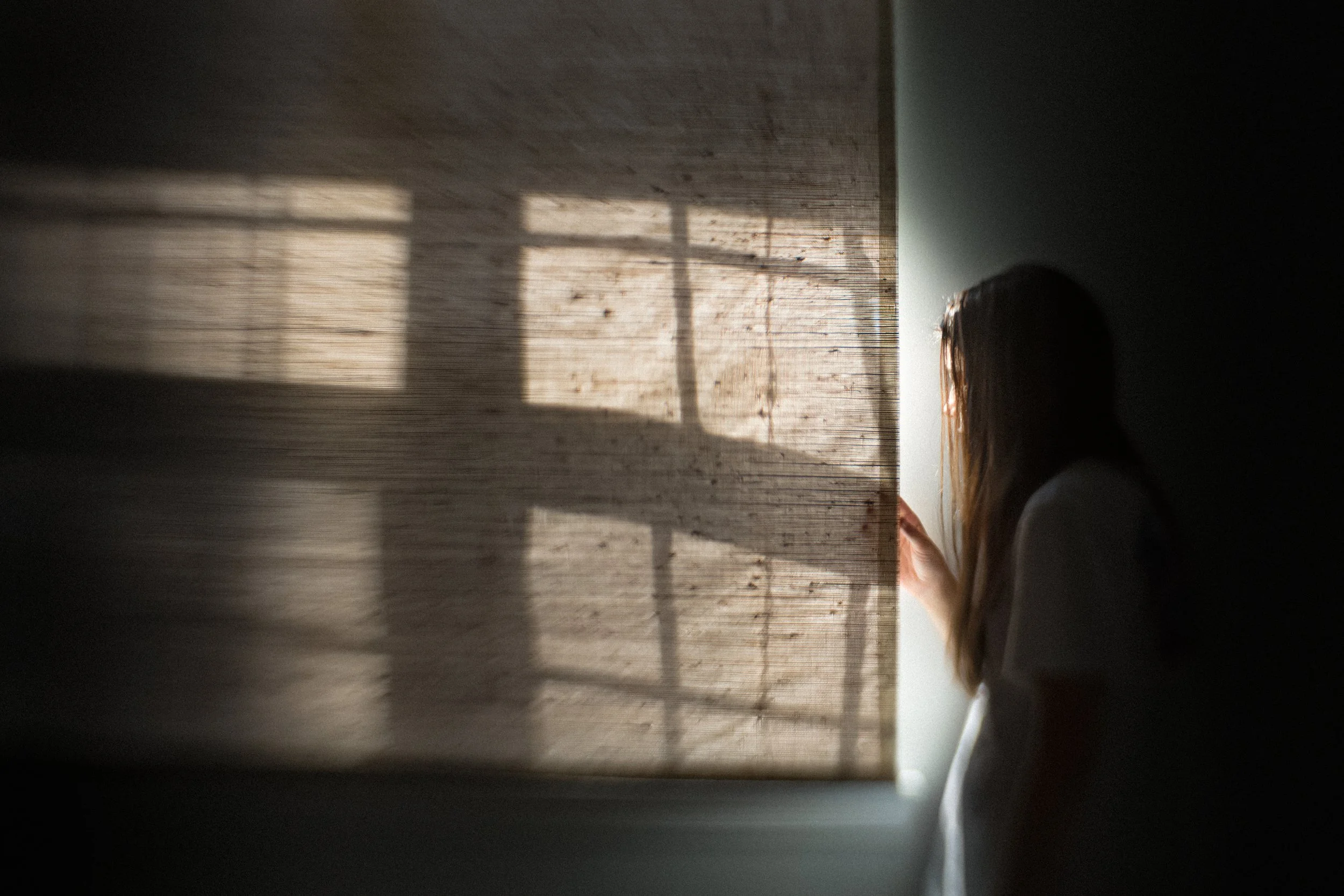 A person with long hair, possibly a woman, is seen from the side, peeking out of a window through a shade, with sunlight casting shadows on the wall.