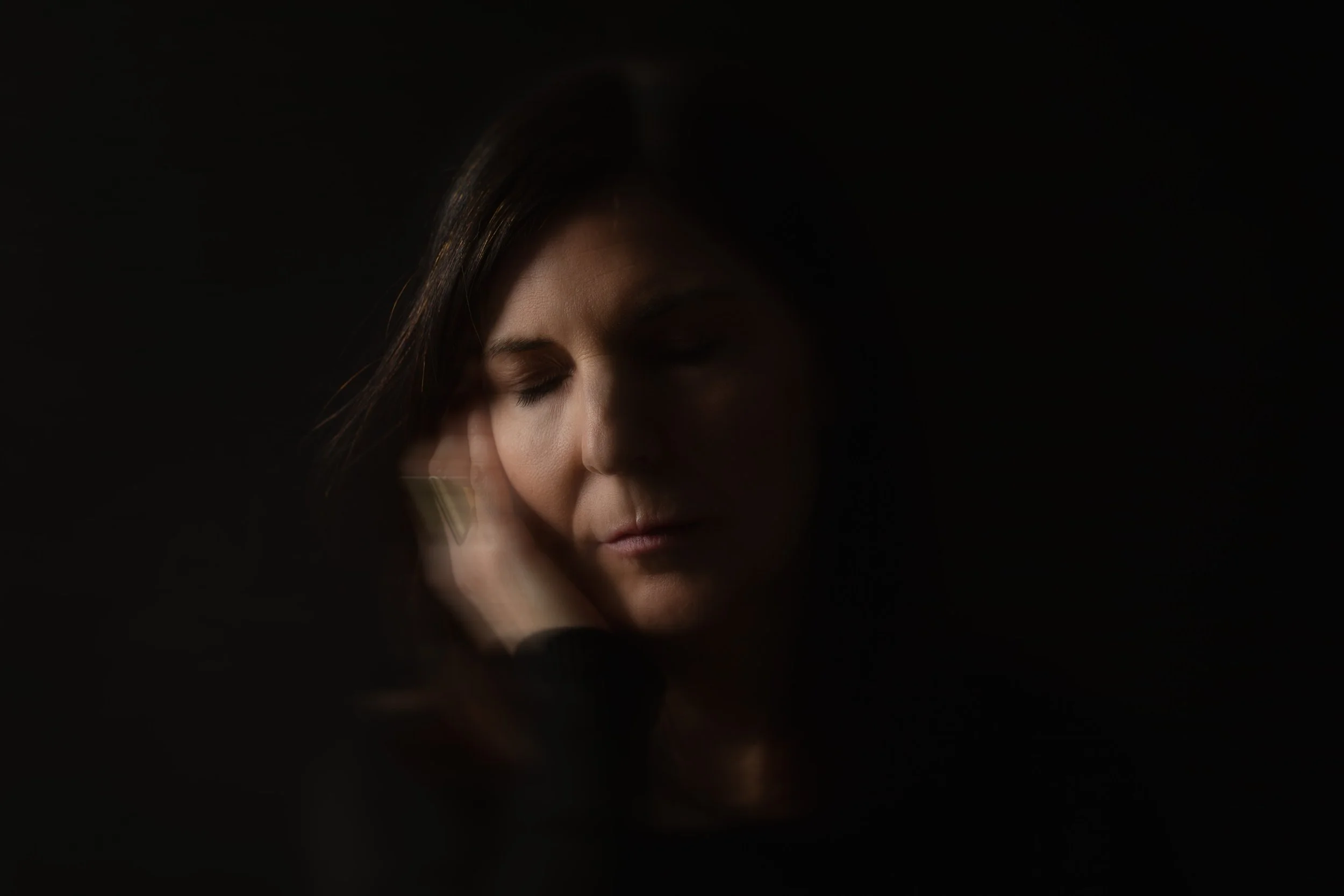 A woman with dark hair and closed eyes, gently holding her face with her hand, set against a dark background.