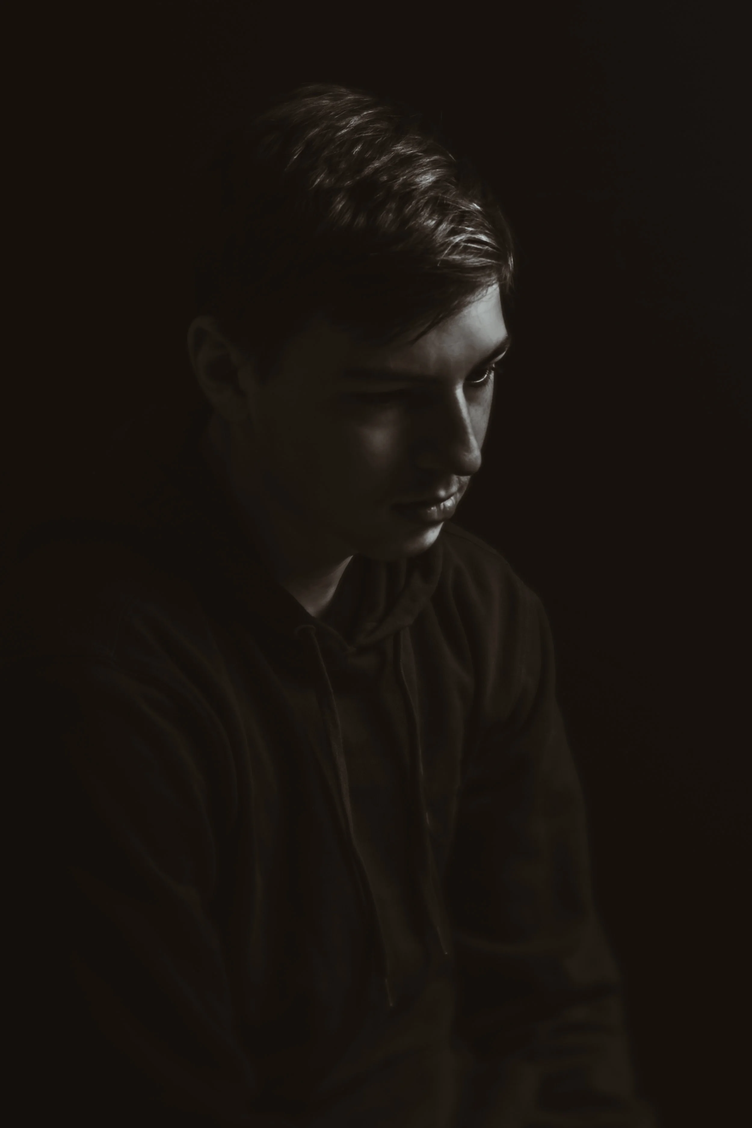 A young person with styled hair looking downward, illuminated by rim light against a dark background, wearing a hooded sweatshirt.