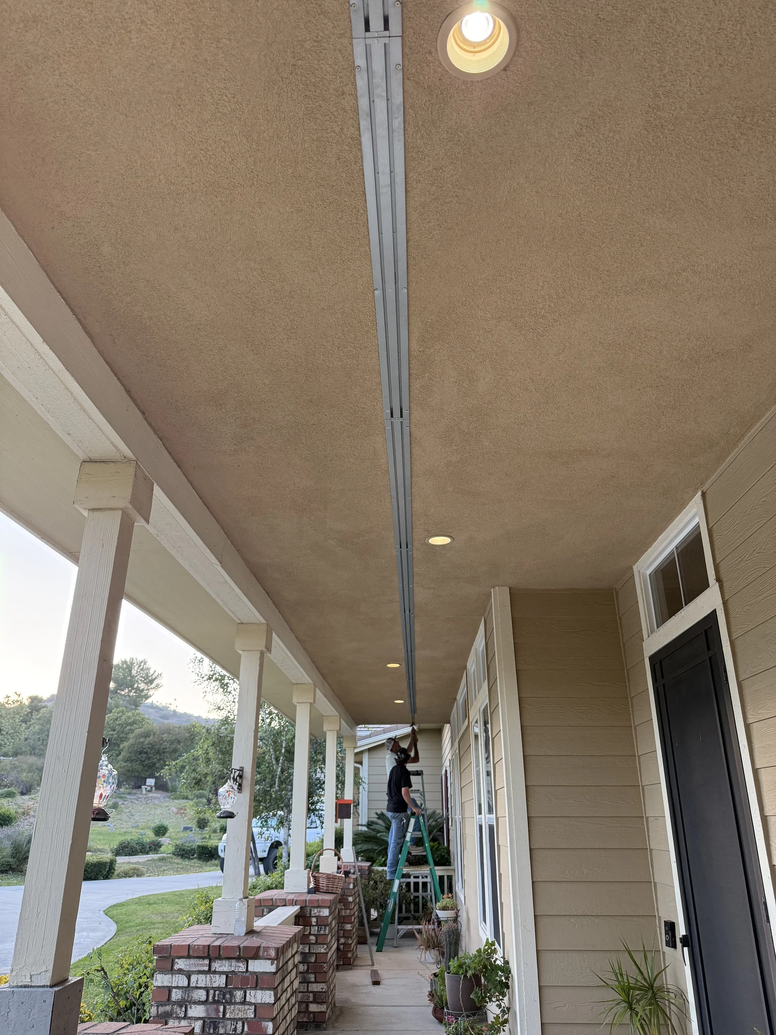 Image of two men installing BrandGuard's linear ember resistant soffit vent in a home's covered stucco patio area at the front of the house