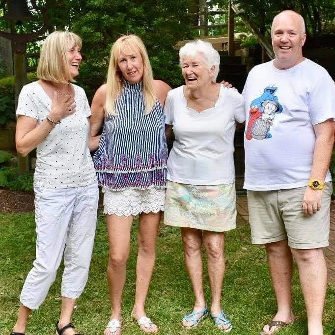 Heather, Sandi, Marjorie and Rob Douglas standing outdoors on grass, smiling and laughing, with trees in the background.