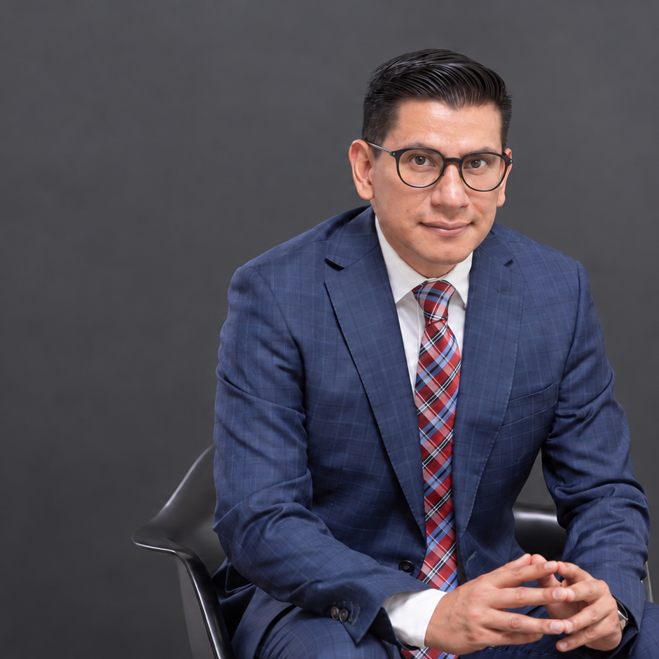 Hombre con gafas, vestido con traje azul y corbata a cuadros, sentado en una silla contra fondo gris.
