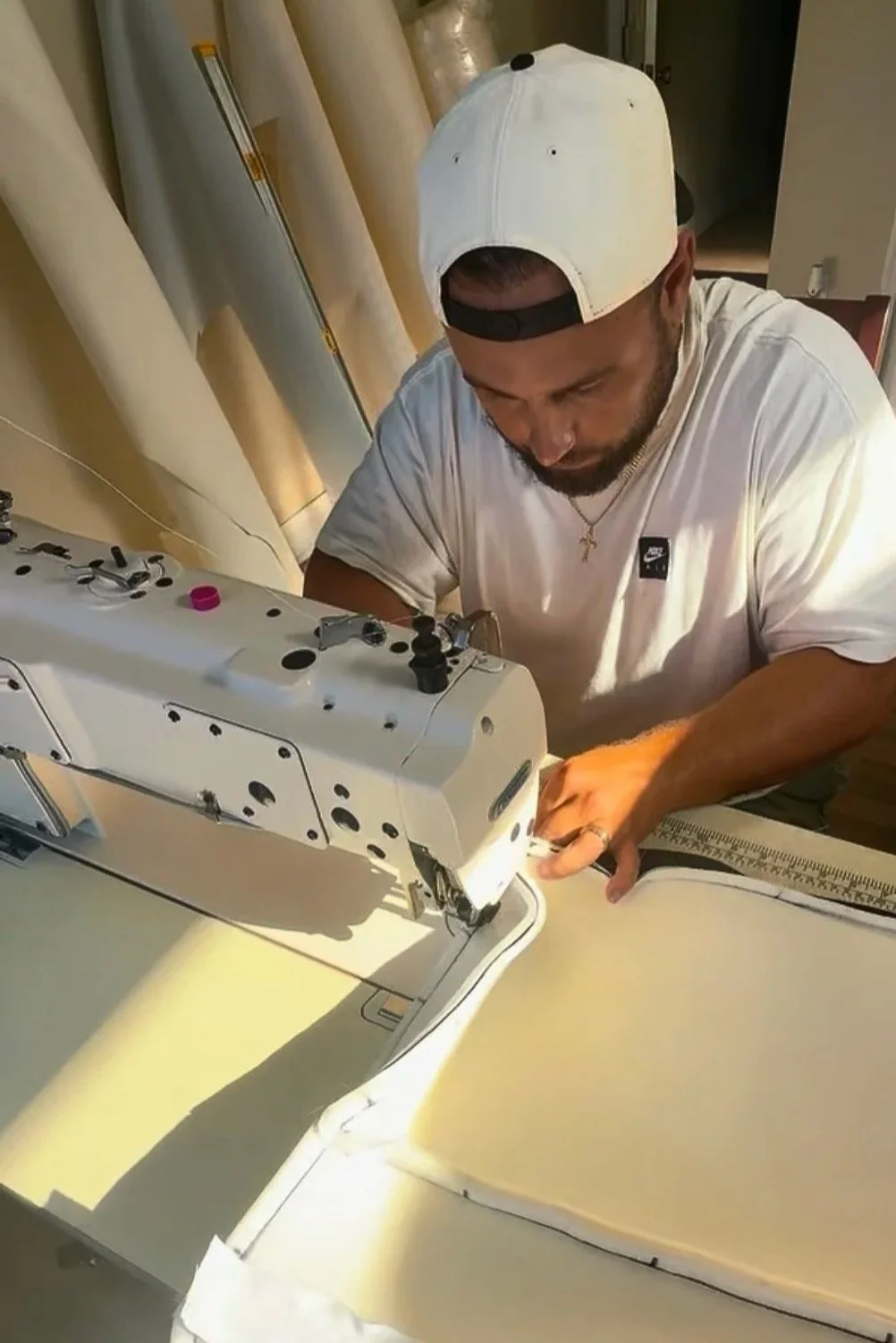 A man wearing a white baseball cap backwards and a white t-shirt sewing fabric using a sewing machine in a well-lit room.