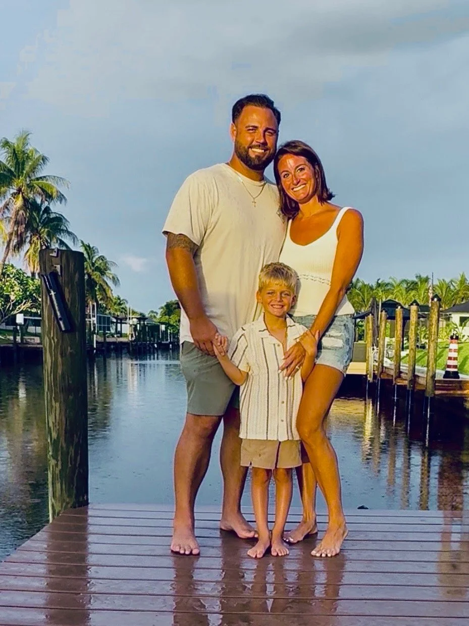 A happy family of three standing on a wooden dock near water with palm trees and a cloudy sky in the background. The father is tall with a beard, wearing a beige t-shirt and shorts. The mother has short dark hair, wearing a white tank top and denim shorts. The young boy has blonde hair, wearing a striped shirt and shorts, and is smiling.