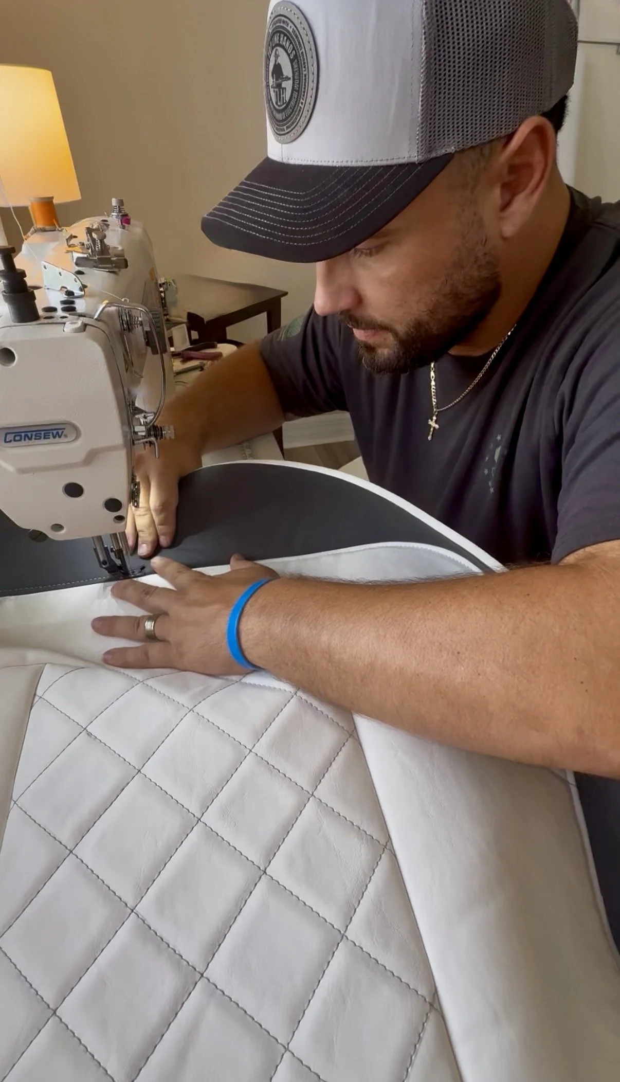 A man operating a sewing machine, working on quilting fabric in a room with a beige wall and a side table with various items, wearing a gray and black cap, a black t-shirt, a silver cross necklace, a blue wristband, and a wedding ring.