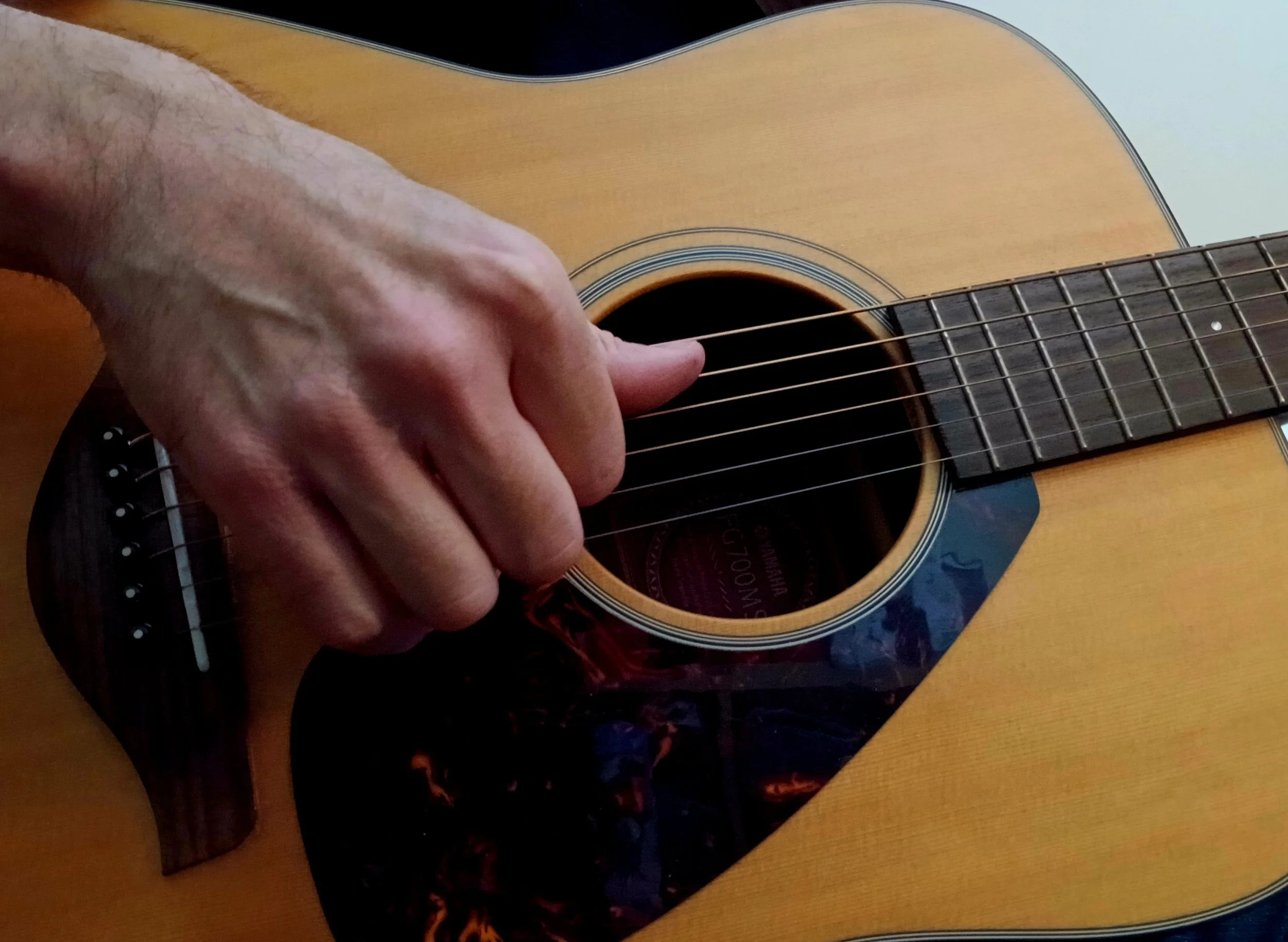 Close-up of someone playing an acoustic guitar, showing their left hand pressing down on the strings on the fretboard.