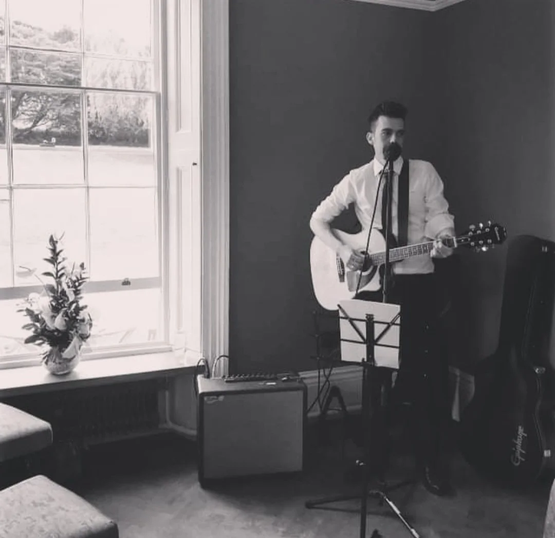 Man with dark hair and a mustache playing an acoustic guitar and singing into a microphone in a room with a large window, a flower vase on the windowsill, and speaker equipment.