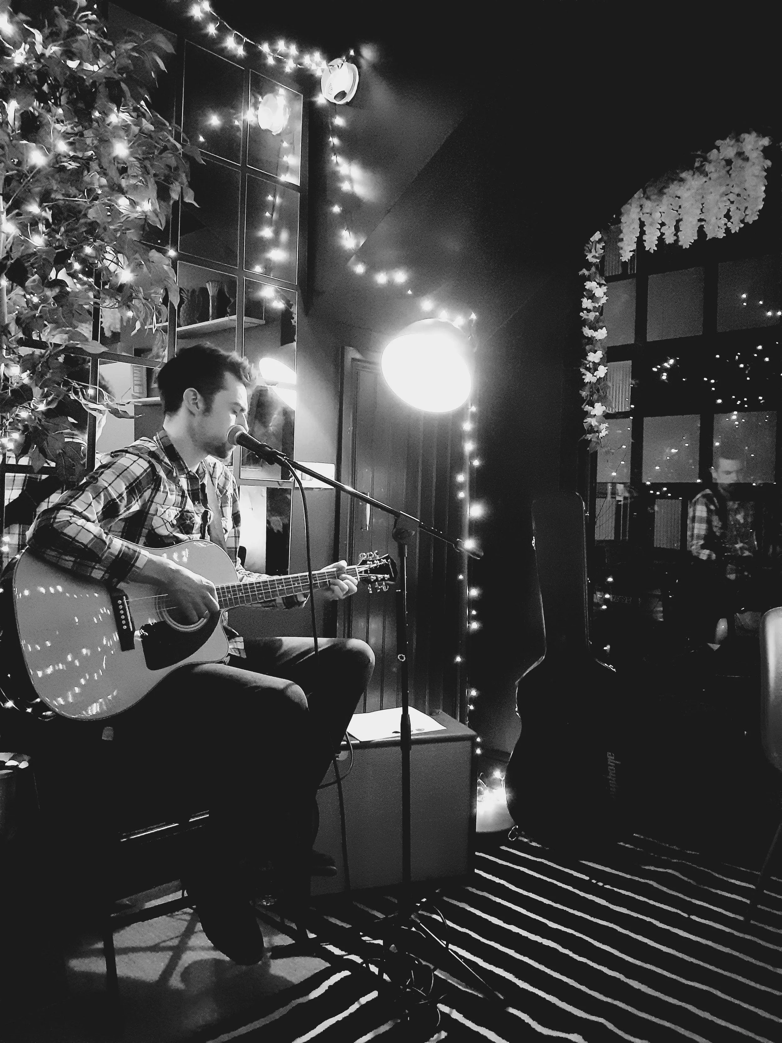 A man in a plaid shirt sitting on a chair, playing an acoustic guitar and singing into a microphone in a cozy room decorated with string lights and floral arrangements.
