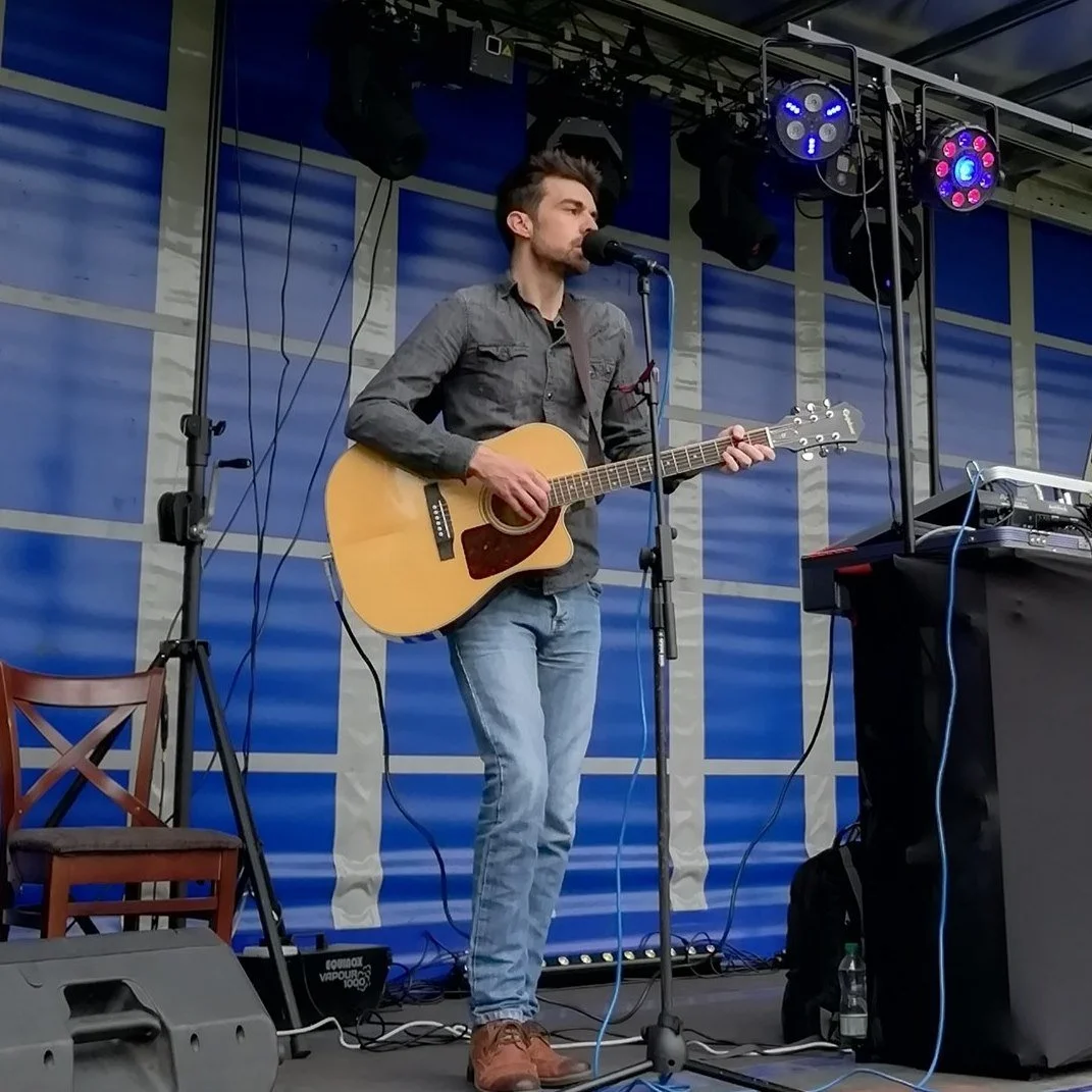 A man plays an acoustic guitar and sings into a microphone on a stage with blue backdrop, lighting equipment, and DJ equipment nearby.
