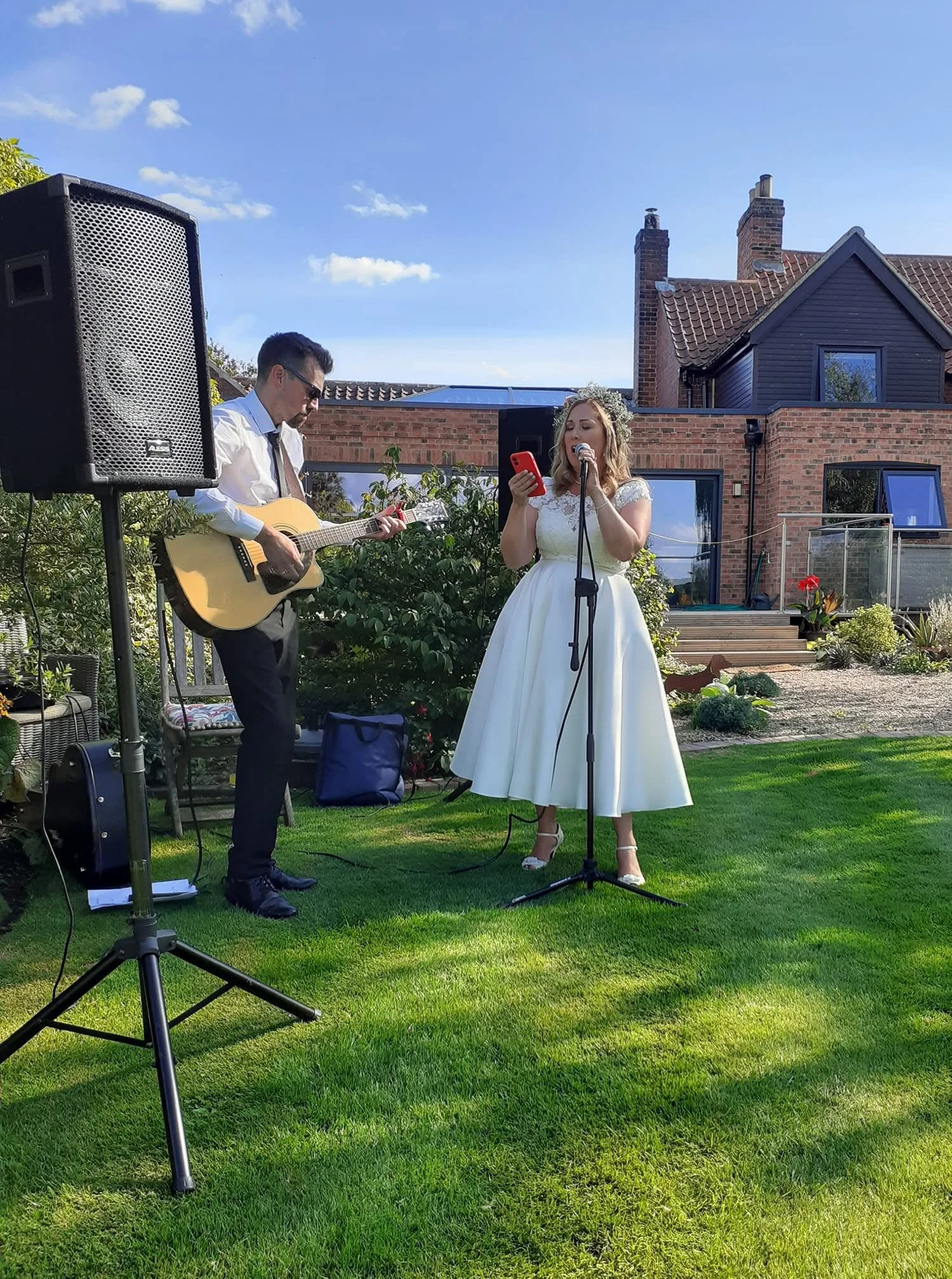 A woman in a white dress singing into a microphone while a man in a white shirt and tie plays a guitar beside her in a backyard with green grass and a house with brick walls and chimneys in the background.