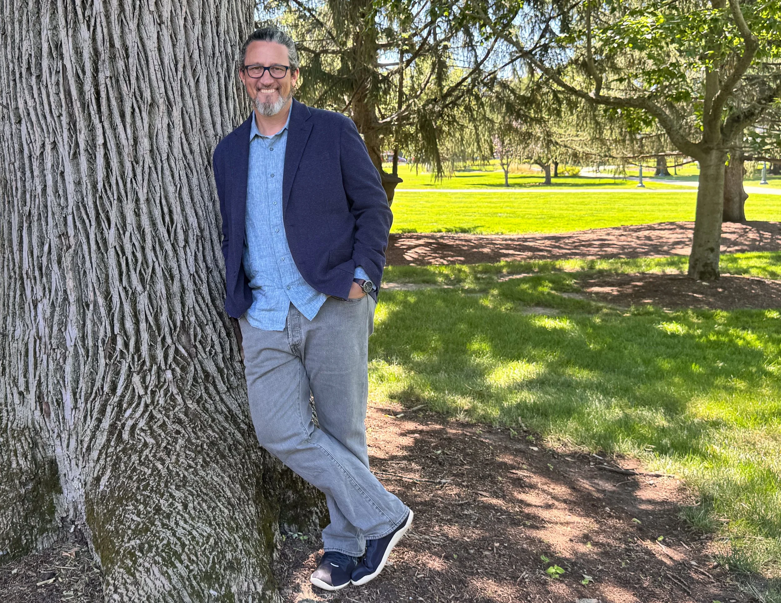 Derek Raynes leaning against a tree outdoors on the University of Vermont campus.