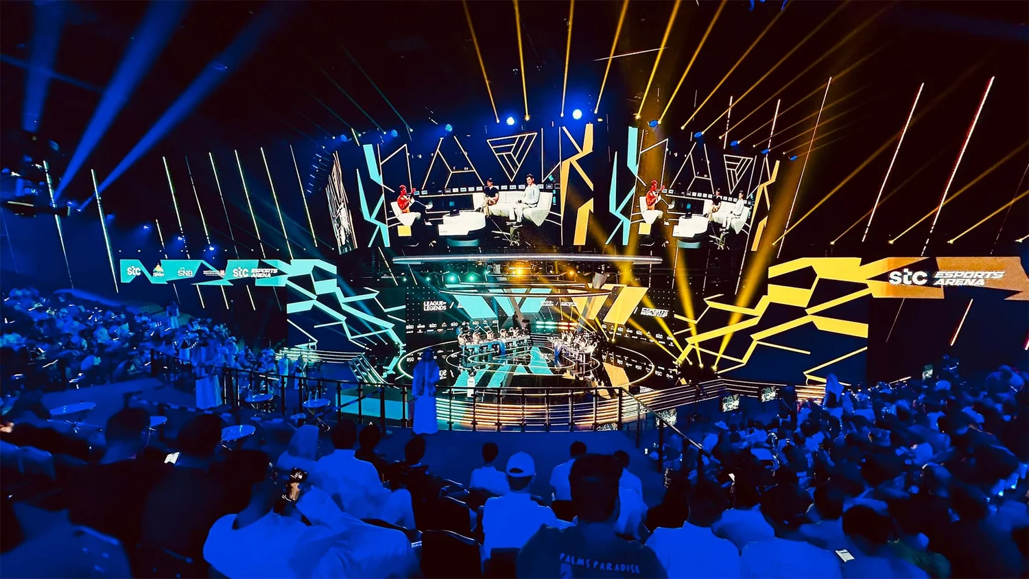 Audience surrounding an esports stage with large screens and blue and yellow lighting.