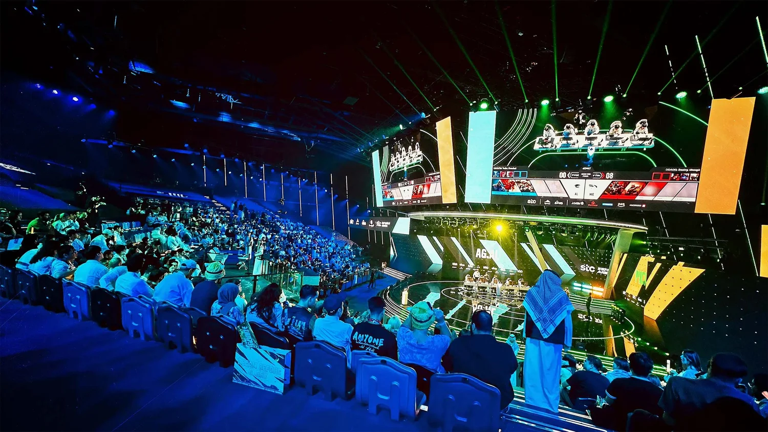 An esports audience watches the stage and player screens under blue and green lighting.