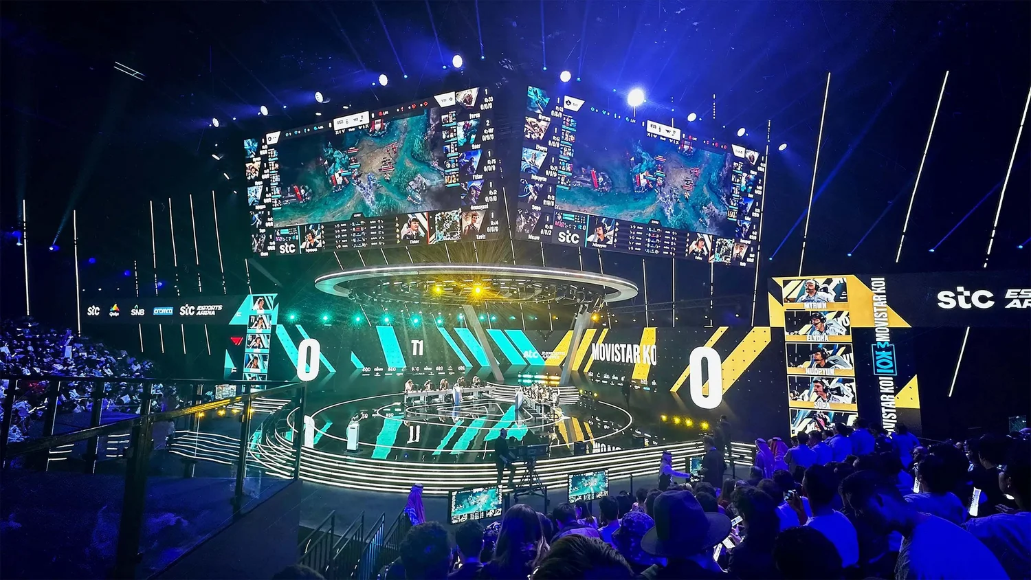 A wide view of an esports arena with a central stage, overhead game screens and blue lighting.